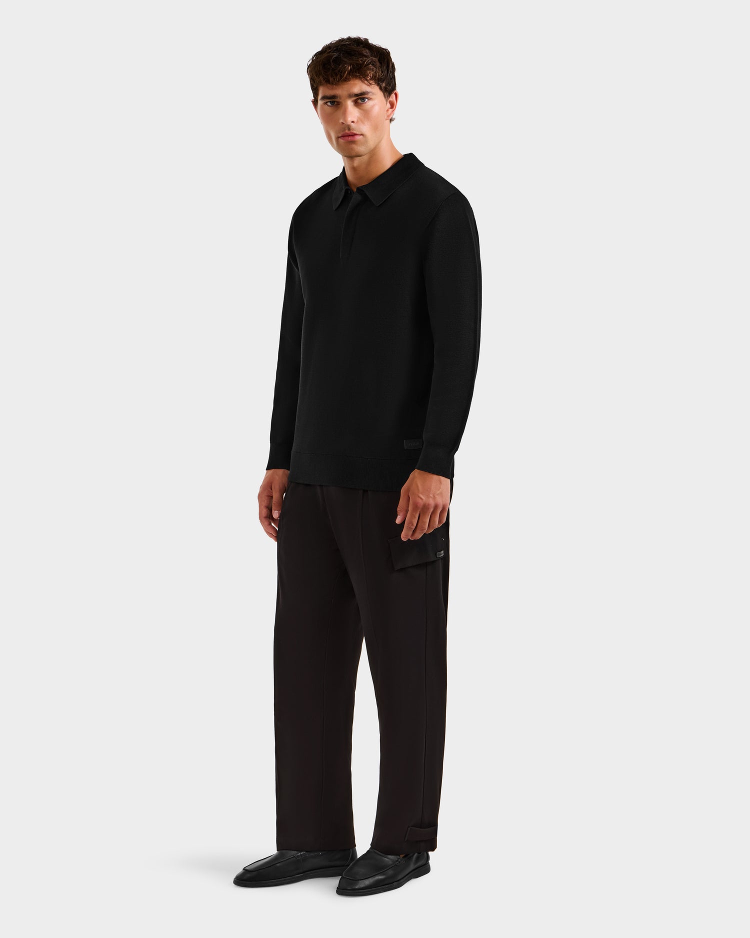 A man stands against a plain background wearing the Rudsak RICHARD - 6524529 Black men’s knit polo sweater, loose dark brown side-pocket pants, and black shoes. He gazes forward with a neutral expression.
