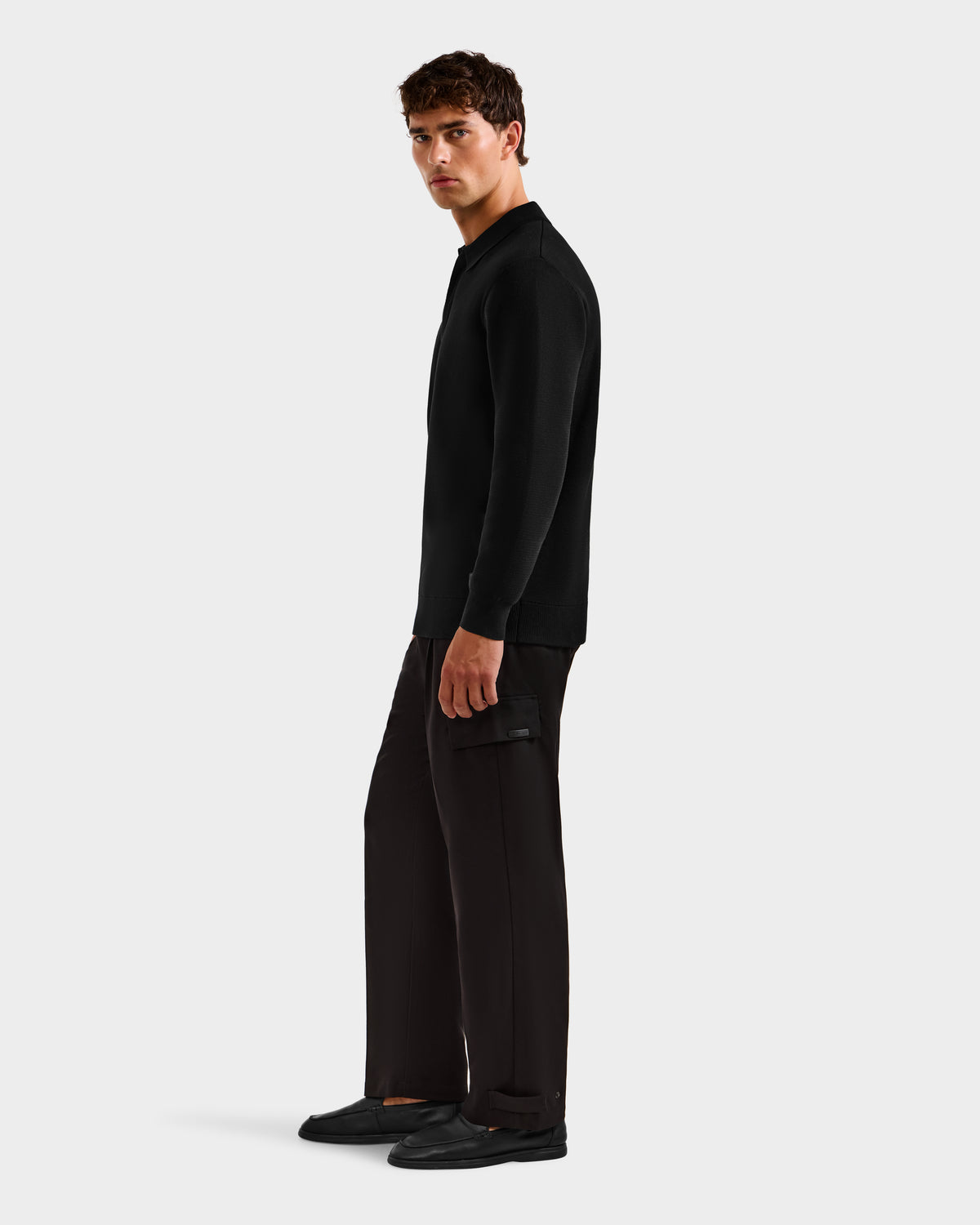 A man stands slightly to the side, wearing the RICHARD - 6524529 Black men's knit polo sweater by Rudsak, paired with dark pants and black shoes against a plain light background.