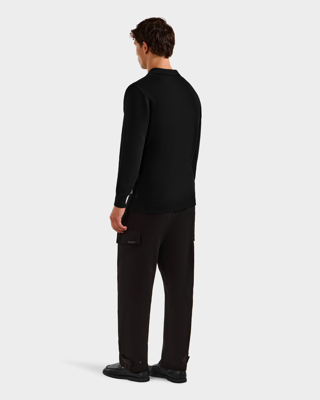A man faces away, wearing the Rudsak RICHARD - 6524529 Black sweater, loose dark pocketed pants, and black shoes, set against a plain light background.