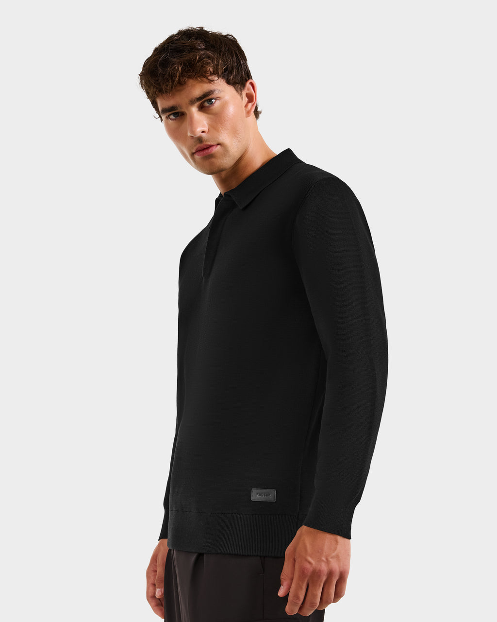 A young man with short brown hair models the RICHARD - 6524529 Black knit polo sweater by Rudsak with dark pants, standing against a plain light gray background and gazing slightly to the side.