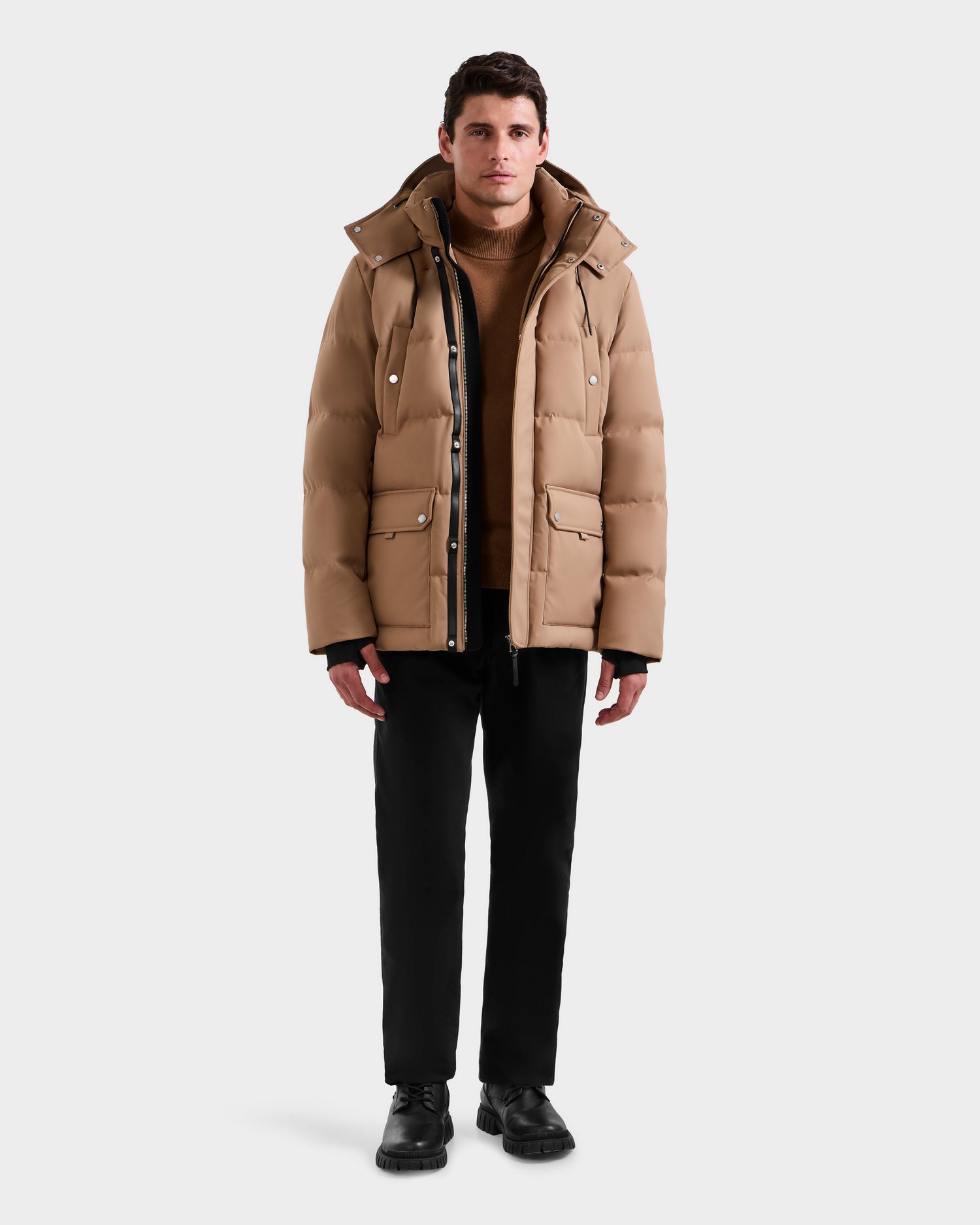 A man faces forward against a white background, modeling the Rudsak RICKY - M123502 Sand men's quilted down parka over a brown sweater, with black pants and shoes, highlighting stylish winter fashion.
