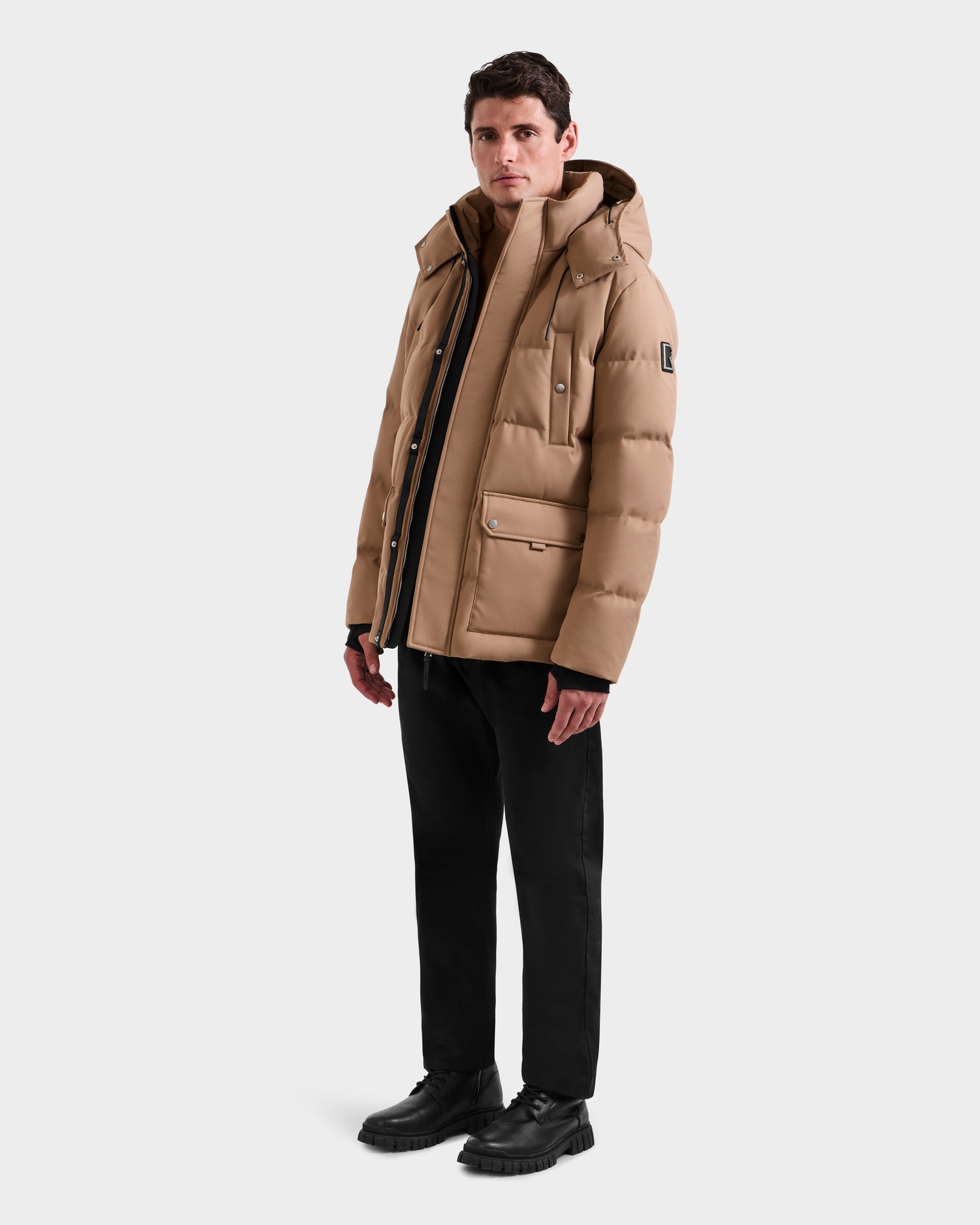 A man stands slightly turned, wearing the Rudsak RICKY - M123502 Sand men's quilted down parka with hood, black pants, and shoes against a white background—ideal for winter fashion.