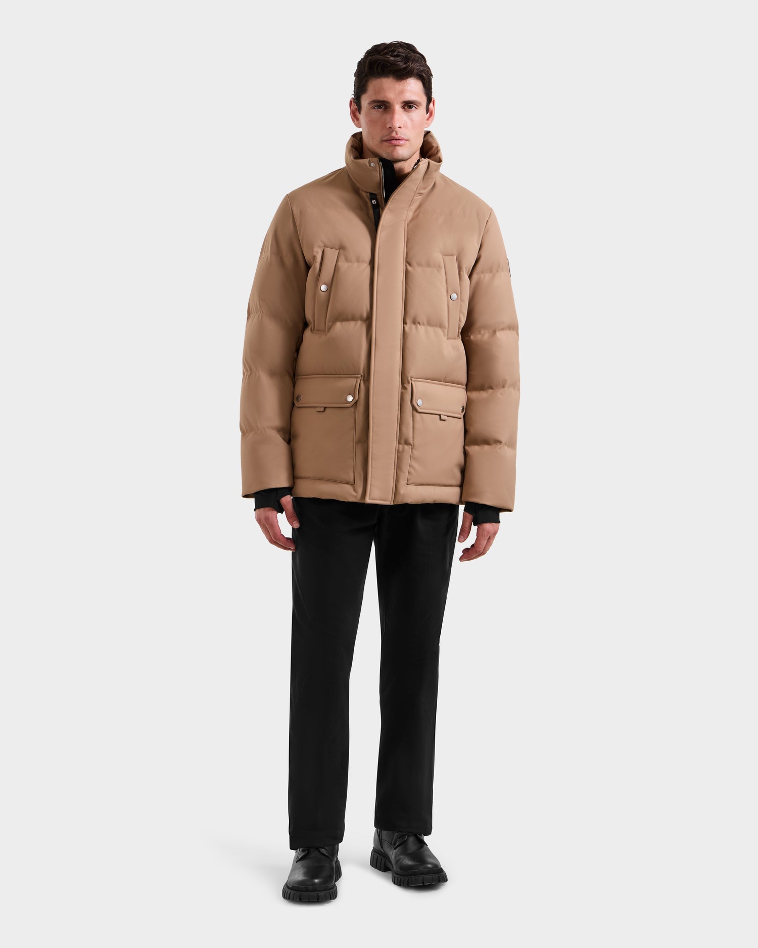 A man stands facing forward in the Rudsak RICKY - M123502 Sand parka, paired with black pants and shoes, embodying modern winter style against a plain light gray background.