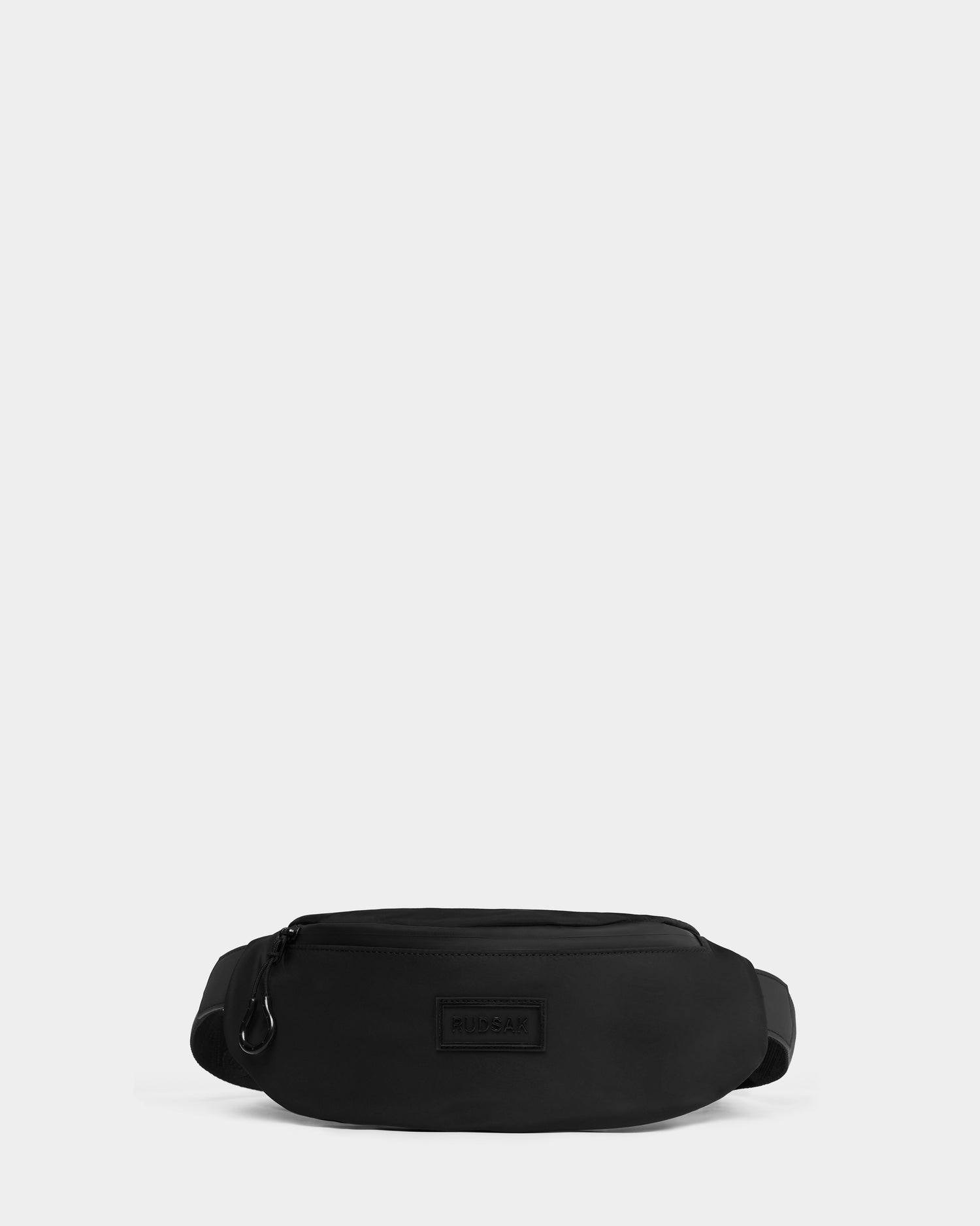 The Rudsak RILO - 9326000 Black waist bag features a minimalist design with a zipper closure, adjustable strap, and a small rectangular logo patch on the front, shown against a plain white background.