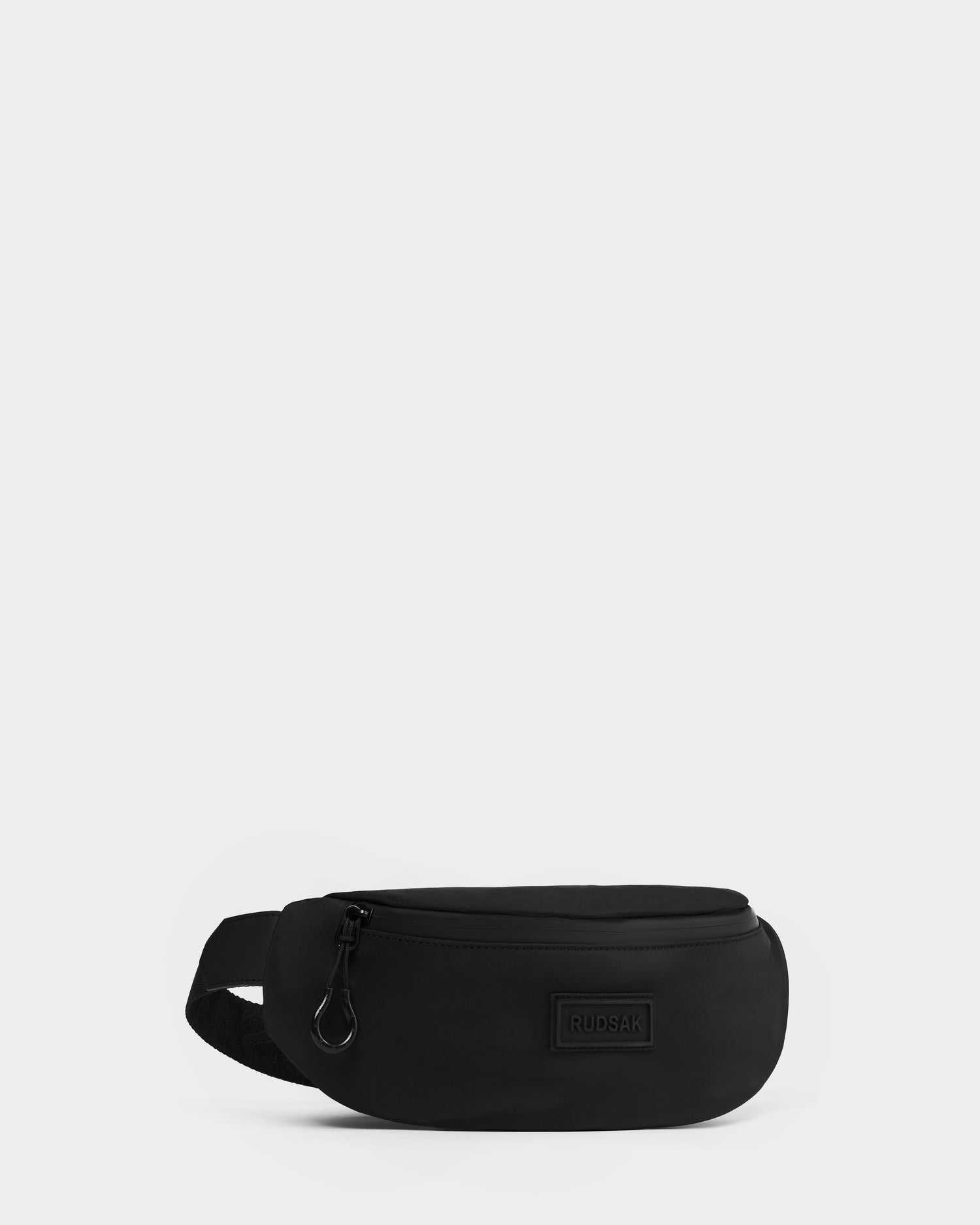 The RILO - 9326000 Black waist bag by Rudsak features a zippered main compartment and adjustable strap, shown against a light gray background. A small rectangular patch with the brand name Rudsak is on the front.