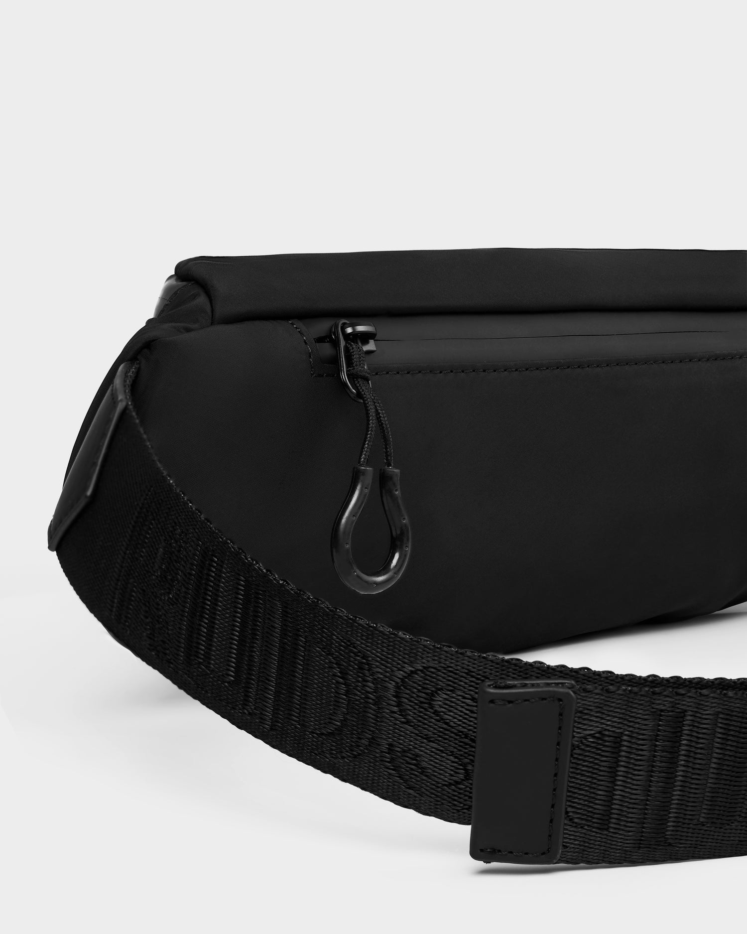 Close-up of the Rudsak RILO - 9326000 Black crossbody or belt bag, with a zippered pocket and wide embossed strap. The sleek, minimalist design stands out against a plain background.