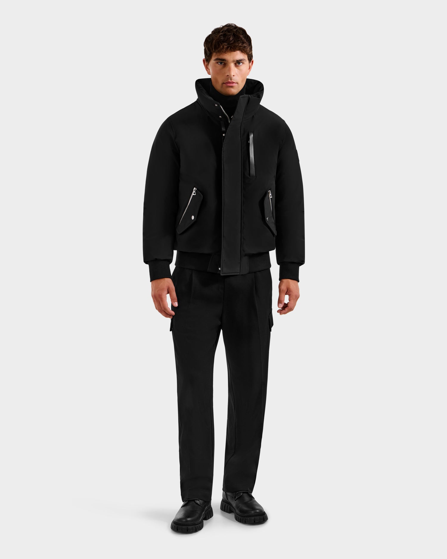 A man faces forward in a Rudsak ROCKY - 6125682 Black recycled nylon winter bomber with zippered pockets, paired with black pants and shoes. The background is plain white.