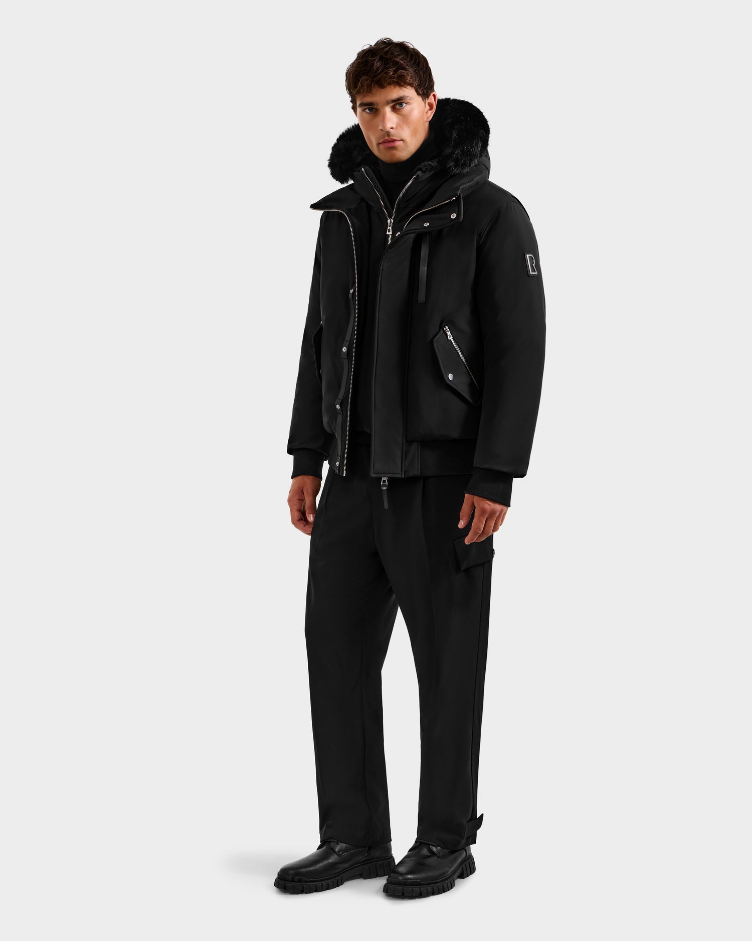 A man stands facing forward in the Rudsak ROCKY - 6125682 Black recycled nylon winter bomber with a fur-lined hood, paired with black pants and shoes, against a plain light background.