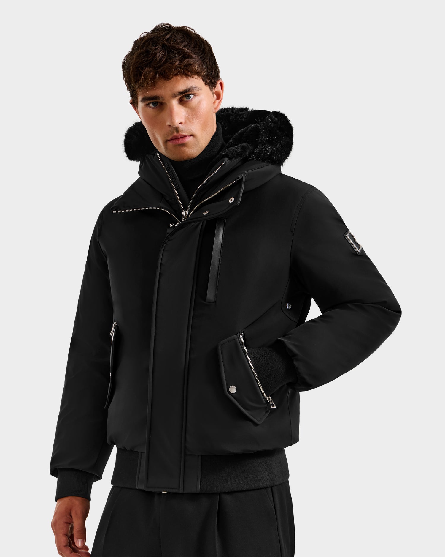 A man in the Rudsak ROCKY - 6125682 Black recycled nylon winter bomber jacket with zipper and snap buttons, black pants, and a turtleneck stands against a plain light background.