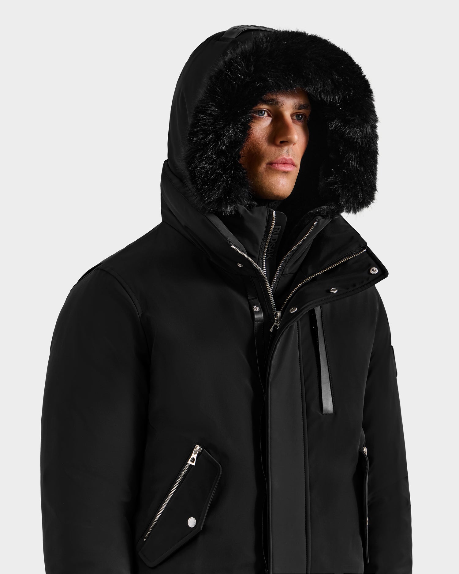 A person wears the Rudsak ROCKY - 6125682 Black recycled nylon winter bomber with a fur-lined hood, partially zipped, standing against a plain light background.