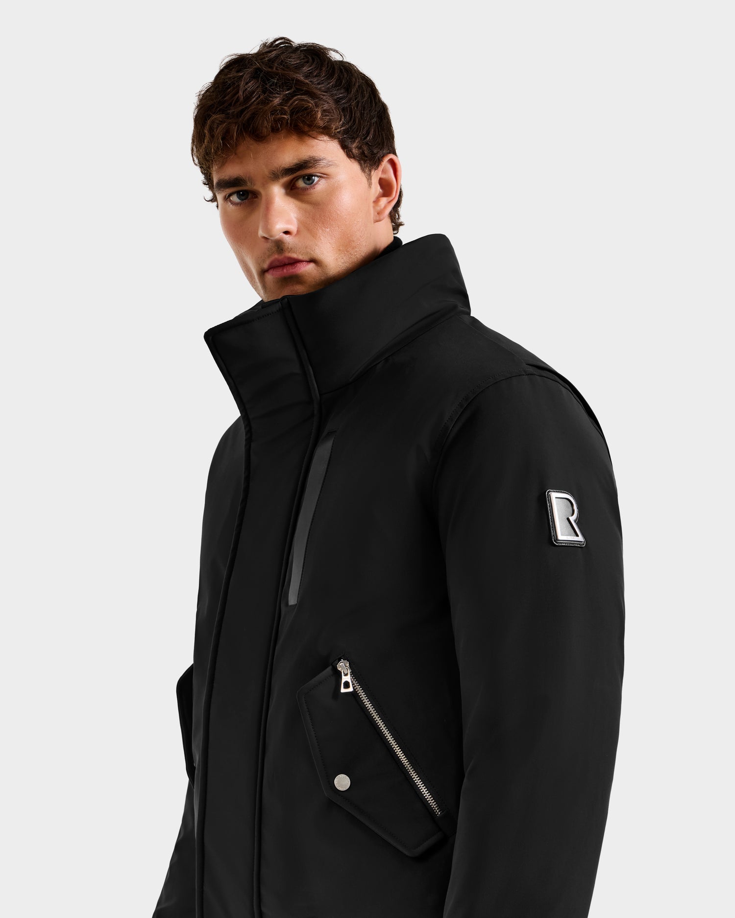 A young man with short brown hair wears the Rudsak ROCKY - 6125682 Black recycled nylon winter bomber with a high collar, zippered pockets, and an “R” logo patch on the left sleeve, looking slightly to the side against a plain light gray background.