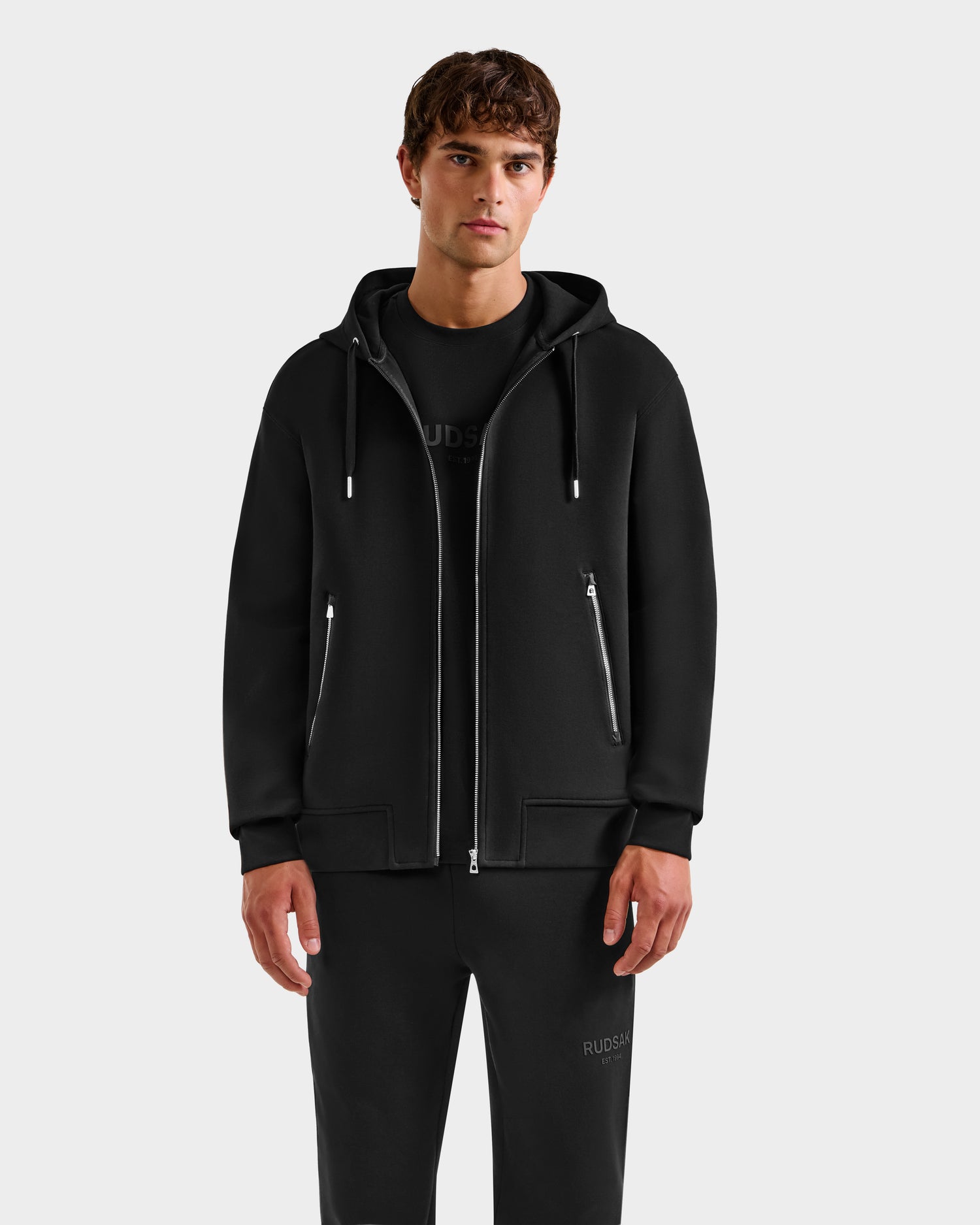 A man faces forward in a Rudsak ROGER - U523507 Black M hoodie with front pockets and matching black pants, showcasing urban style against a plain light gray background.