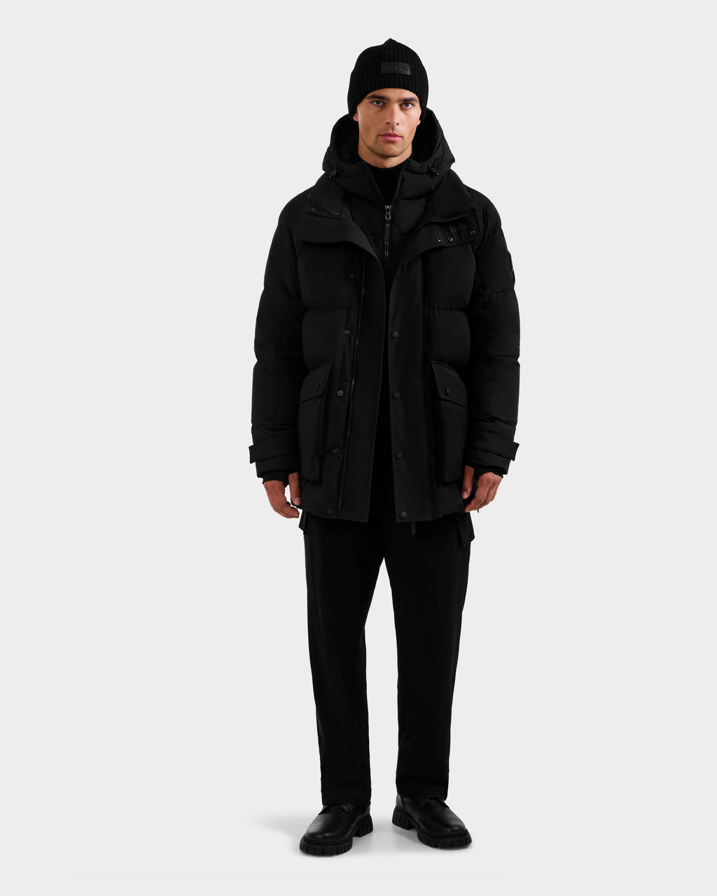 A man faces forward wearing the Rudsak ROKO - 6124510 Black men's quilted down parka with RDS-certified insulation, a black beanie, black pants, and shoes, standing against a plain light gray background.