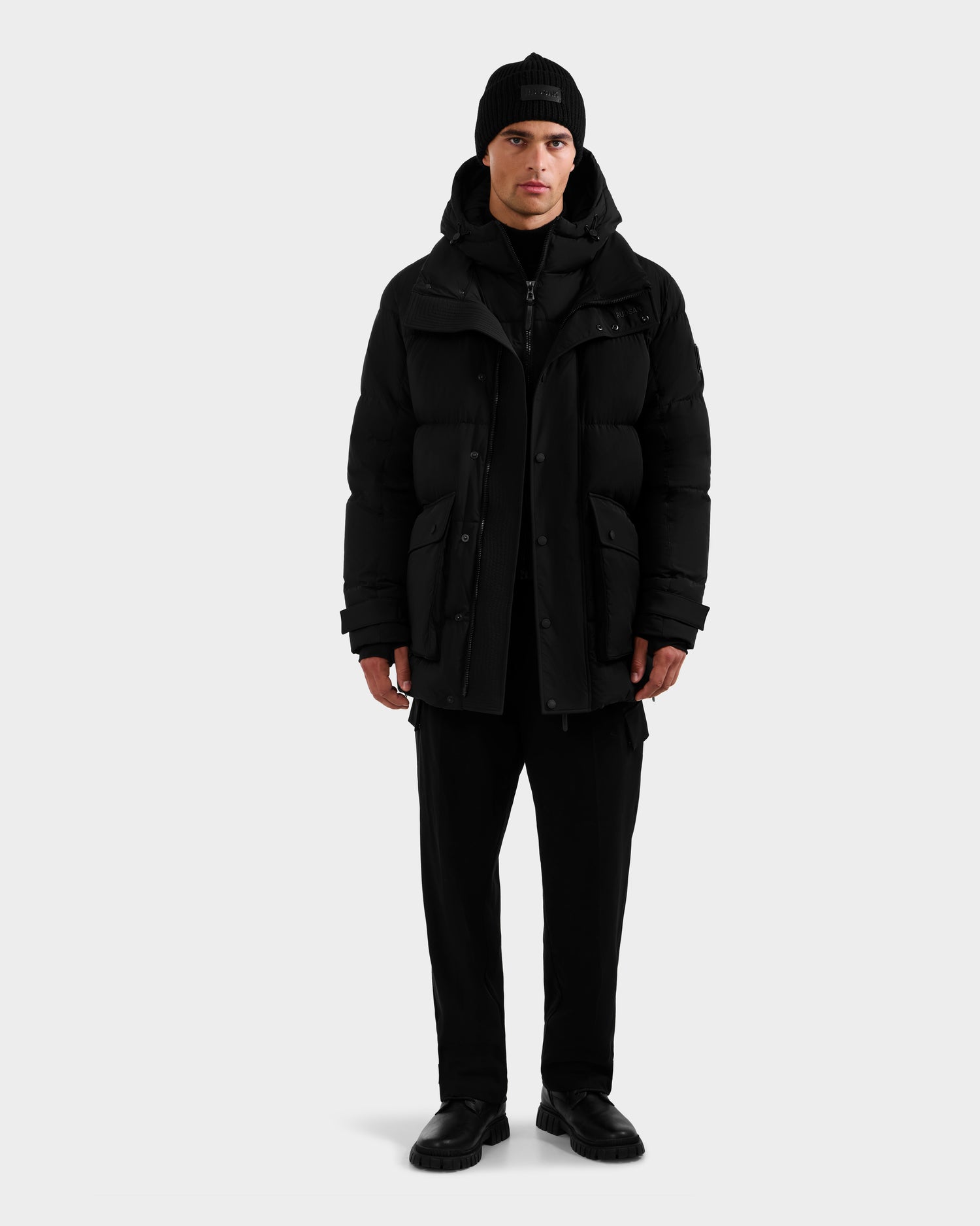 A man faces forward wearing the Rudsak ROKO - 6124510 Black men's quilted down parka with RDS-certified insulation, a black beanie, black pants, and shoes, standing against a plain light gray background.