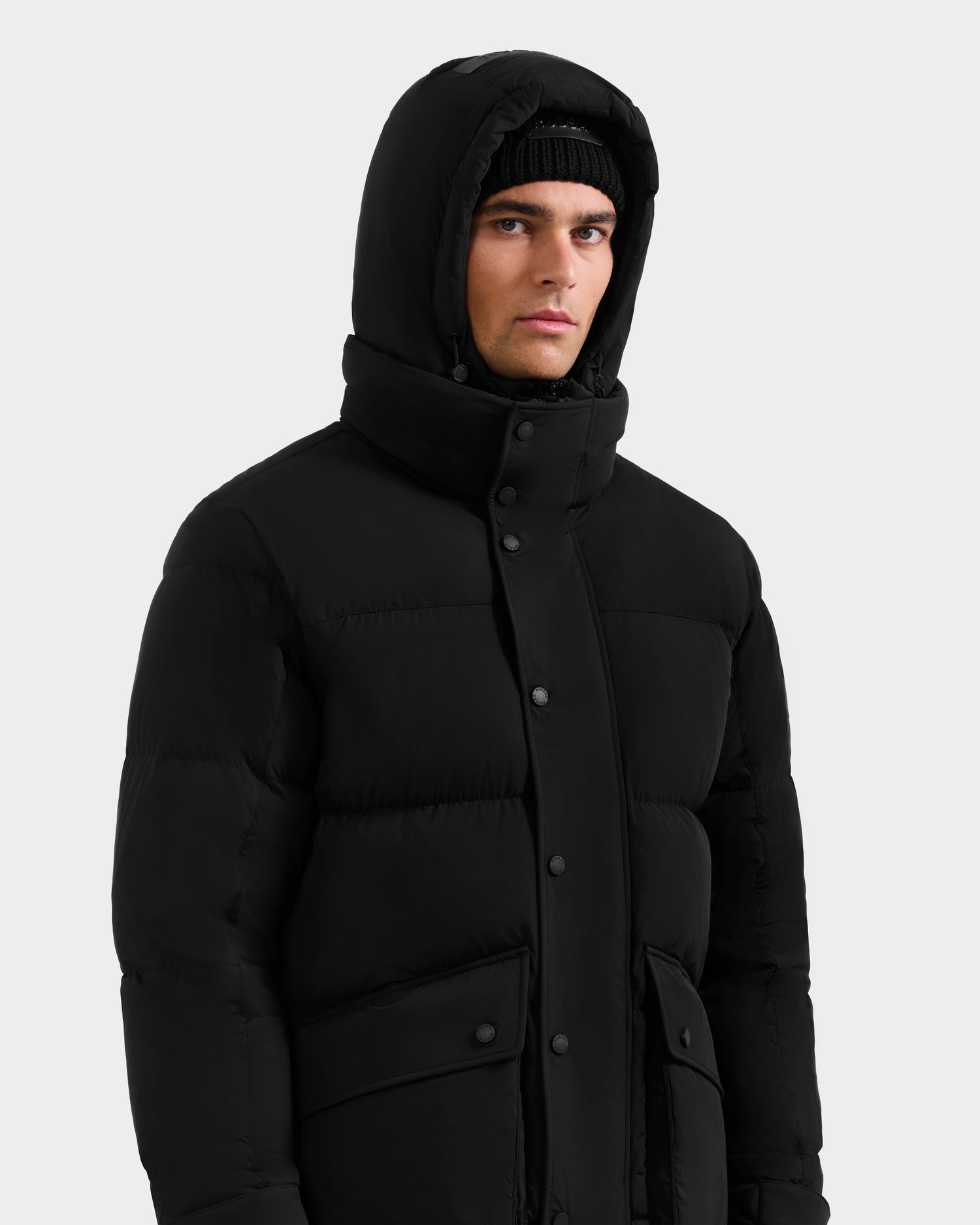 A person wearing the Rudsak ROKO - 6124510 Black men’s quilted down parka with the hood up and a black knit beanie, standing against a plain light gray background.