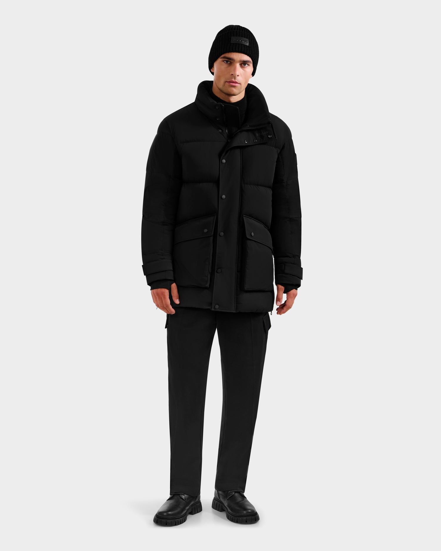 A man stands against a plain background wearing the Rudsak ROKO - 6124510 Black quilted down parka, black beanie, black pants, and black shoes. He has a neutral expression with relaxed hands at his sides.