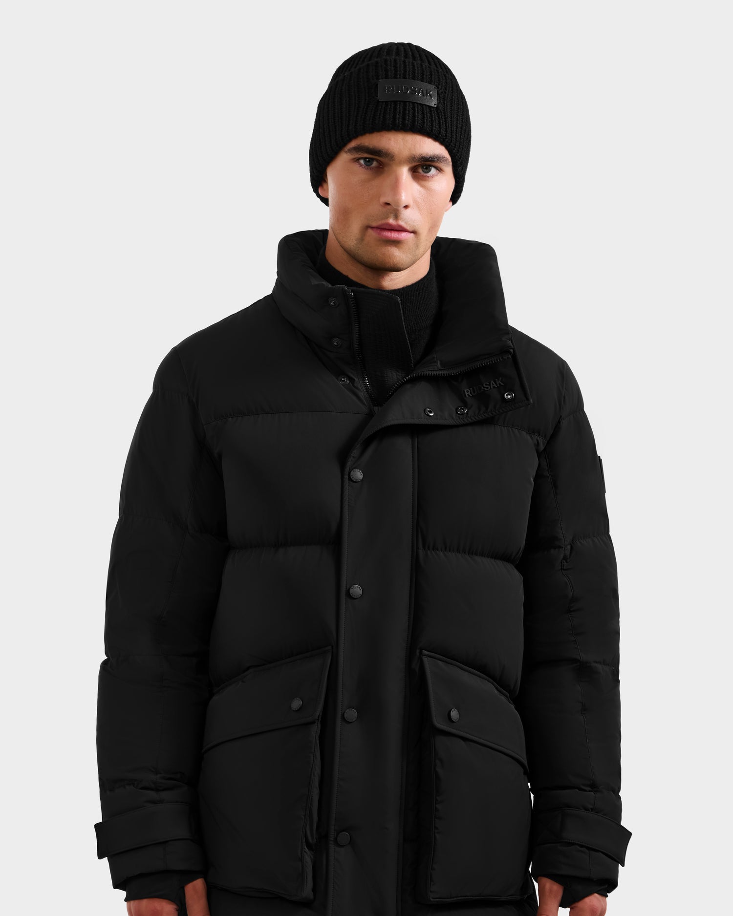 A man wearing the Rudsak ROKO - 6124510 Black quilted down parka with RDS-certified insulation and a black knit beanie stands against a plain light gray background, looking directly at the camera with a neutral expression.