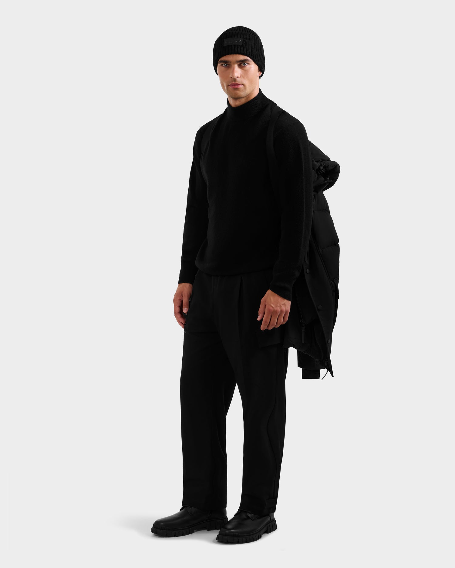 A man in all-black stands against a plain background, holding the Rudsak ROKO - 6124510 Black men’s parka with RDS-certified down over his shoulder, looking forward with a neutral expression.