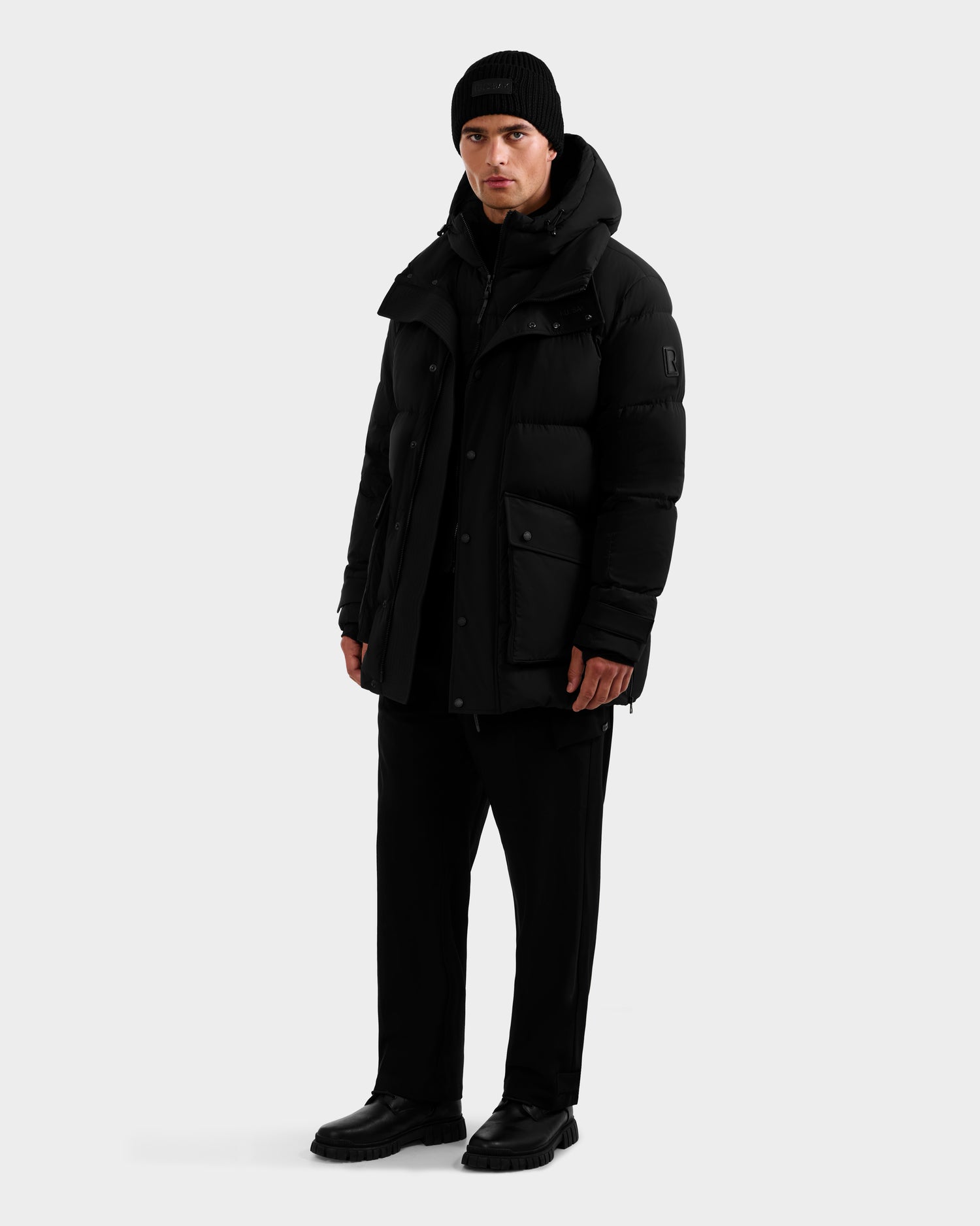 A person stands facing forward in a black Rudsak ROKO - 6124510 men's quilted down parka with RDS-certified insulation, paired with a black knit beanie, pants, and boots, against a plain white background.