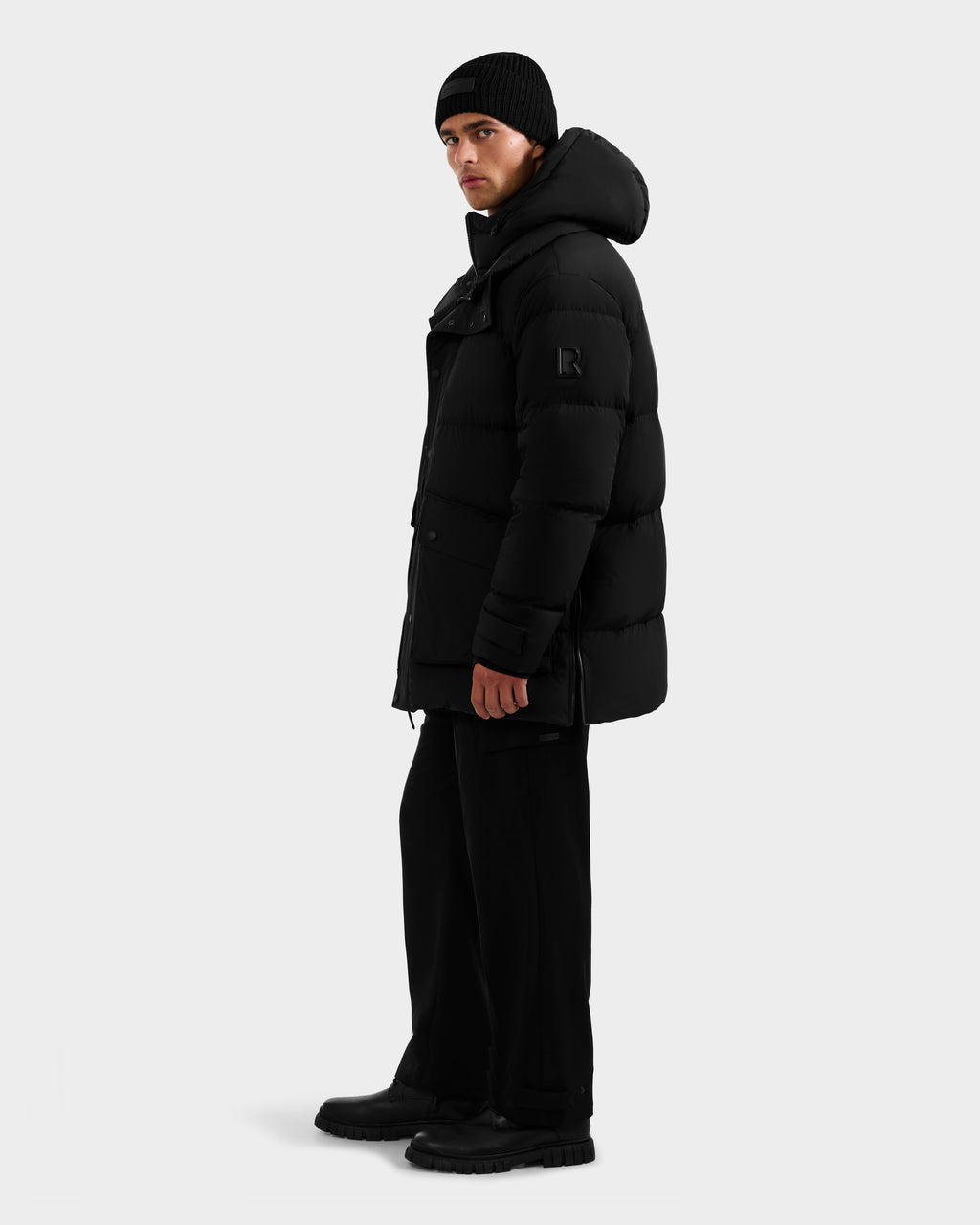 A person wearing the Rudsak ROKO - 6124510 Black men's quilted down parka with a hood, paired with black pants, boots, and a beanie, stands sideways against a plain white background.
