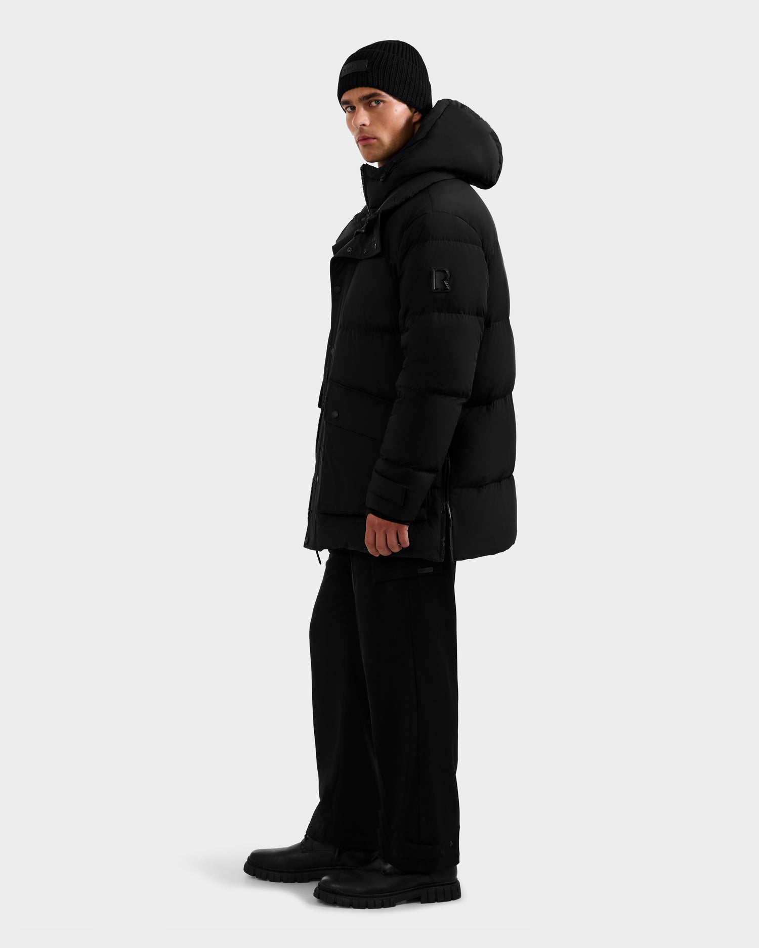 A person wearing the Rudsak ROKO - 6124510 Black men's quilted down parka with a hood, paired with black pants, boots, and a beanie, stands sideways against a plain white background.