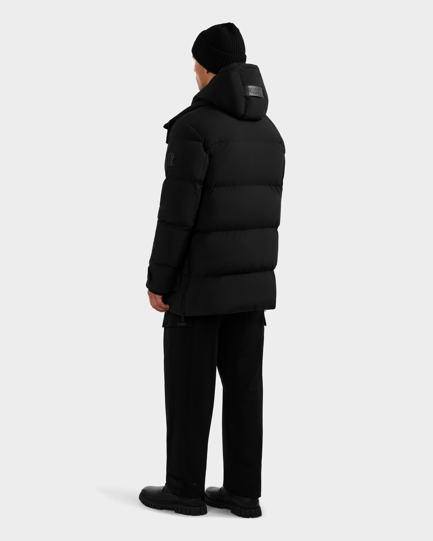 A person wearing the Rudsak ROKO - 6124510 Black men's quilted down parka, paired with black pants, shoes, and a beanie, stands facing away from the camera against a plain light background.
