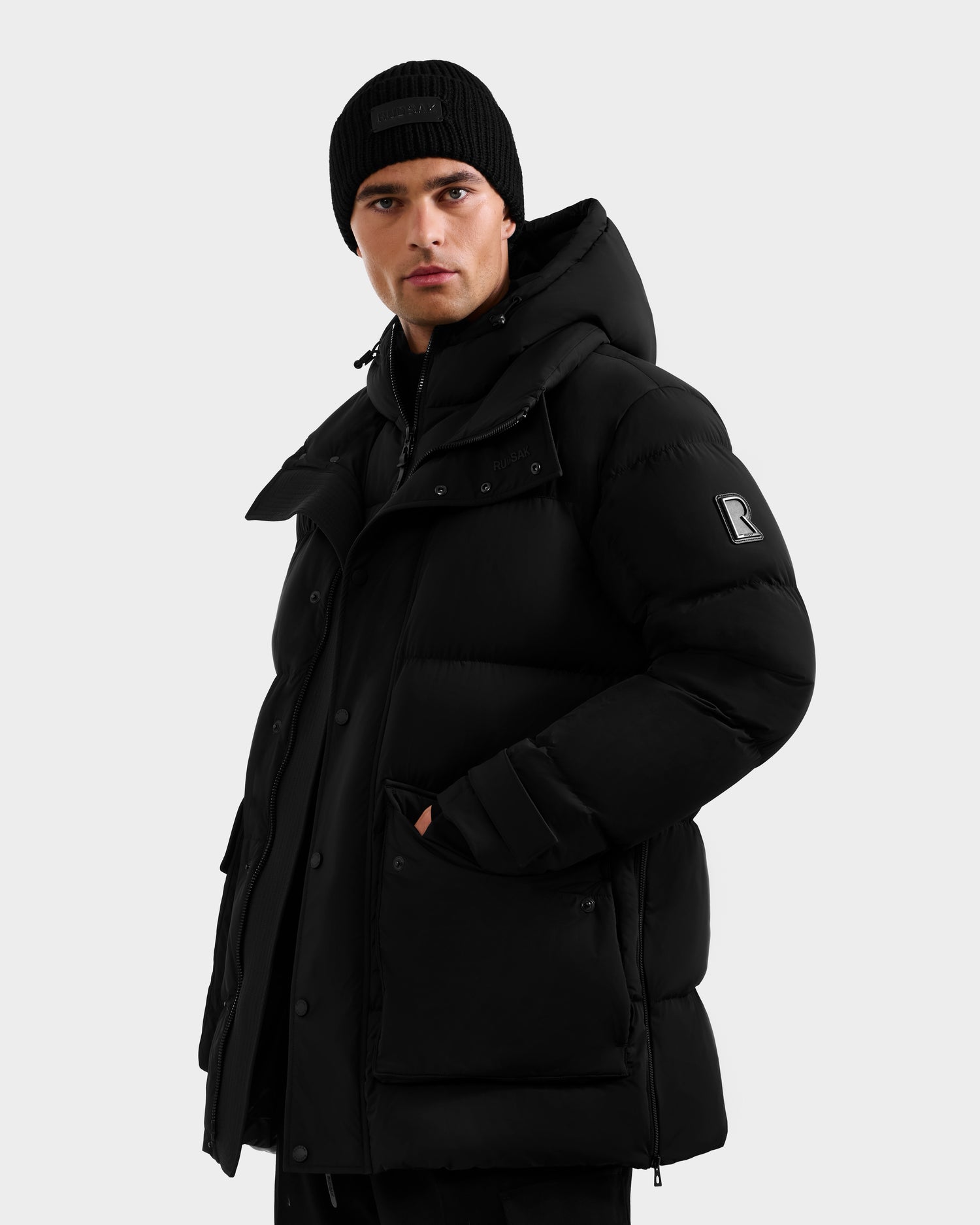 A man in the Rudsak ROKO - 6124510 Black parka with RDS-certified down insulation, large pockets, and a high collar wears a black beanie and looks to the side against a plain light gray background.