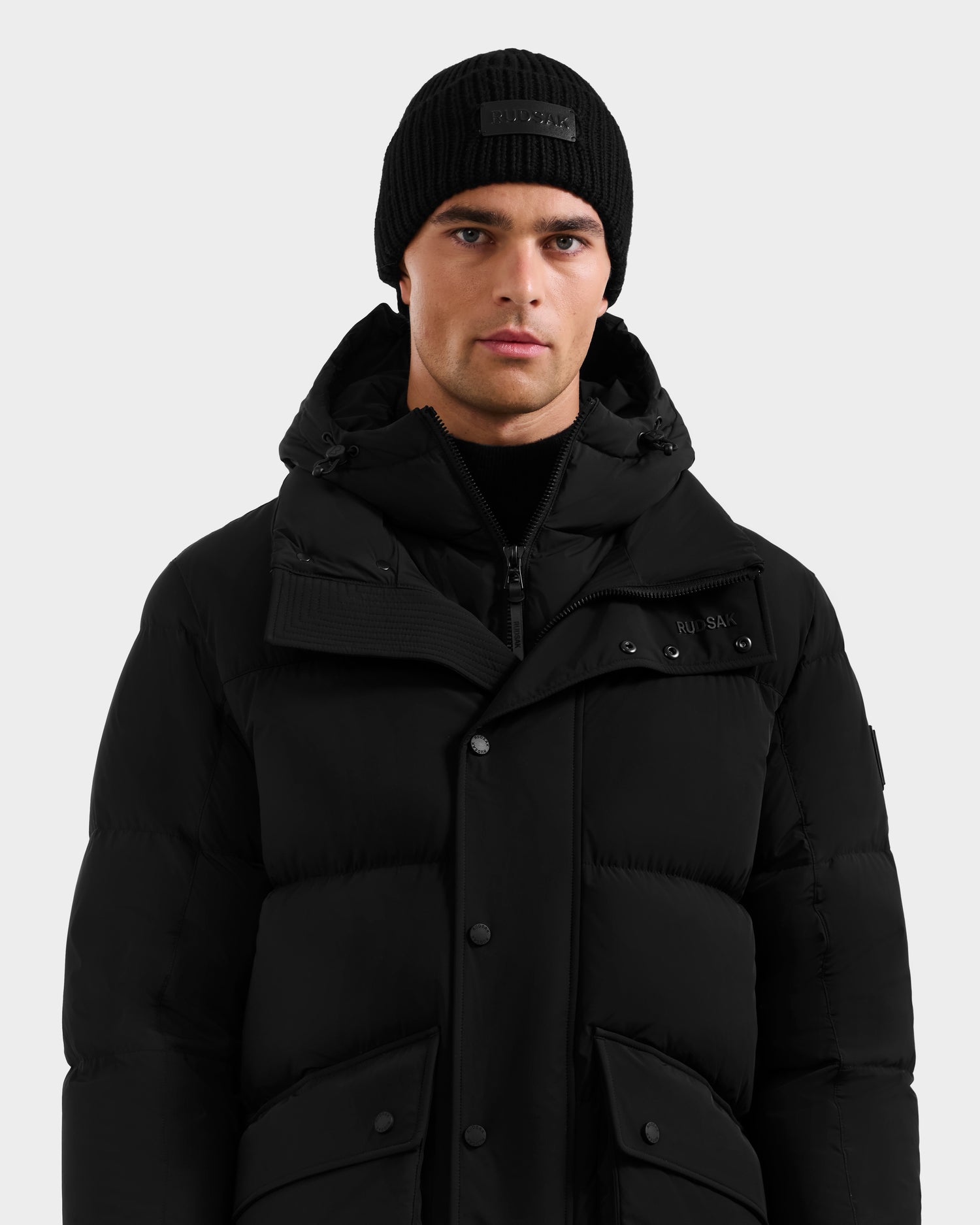 A man wearing the Rudsak ROKO - 6124510 Black quilted down parka with a hood and a black knit beanie stands against a plain light gray background, looking directly at the camera.