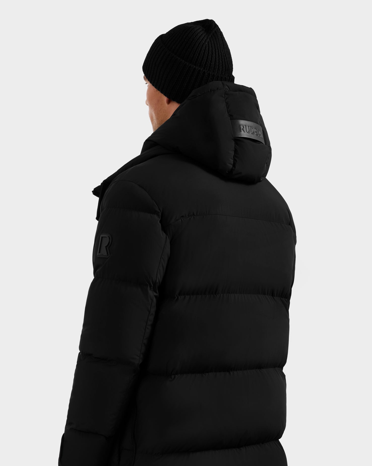 A person is seen from behind in a Rudsak ROKO - 6124510 Black men's quilted down parka with hood and black knit beanie, against a light gray background. The parka features RDS-certified down and a RUDSAK patch on the hood.