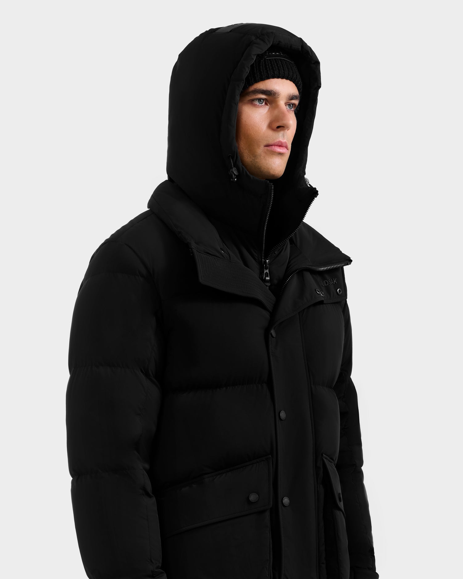 A person wearing the Rudsak ROKO - 6124510 Black men's quilted down parka with the hood up and a black knit beanie, looking slightly to the side against a plain light background.