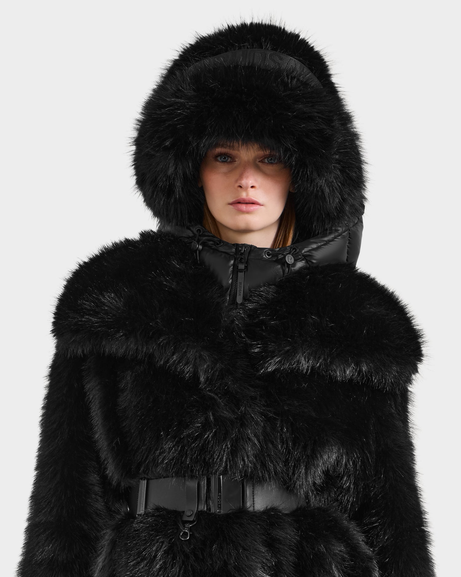 A woman stands facing forward against a plain white background wearing the Rudsak ROMINA - 8425545 Black faux fur coat with hood and belt, paired with a stylish faux fur bucket hat for a cozy-luxe look.