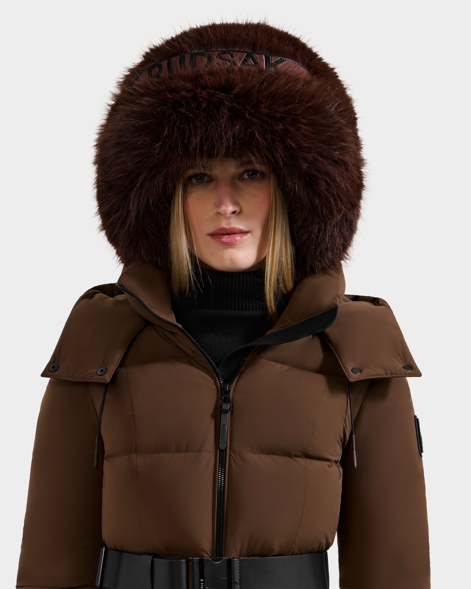 A woman wears the Rudsak ROMINA - 8425545 Espresso puffer jacket with a dark brown fur-trimmed hood and a black turtleneck, standing against a plain light background and showcasing this cozy-luxe winter essential.
