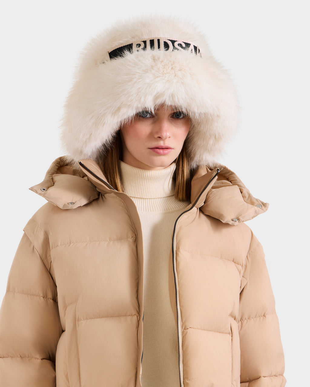 A woman wears the Rudsak ROMINA - 8425545 Vanilla puffer jacket, featuring a large white fur hood and black lettering over a cream turtleneck, standing against a plain light background.