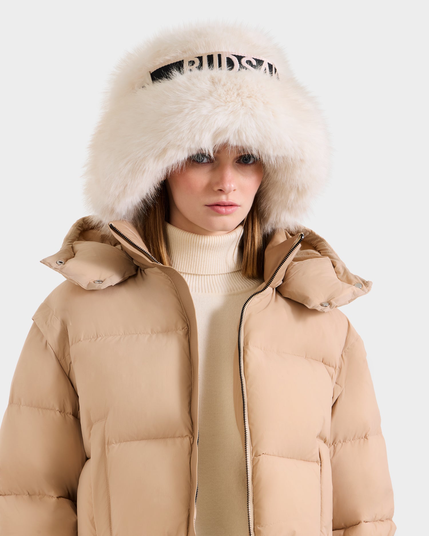 A woman wears the Rudsak ROMINA - 8425545 Vanilla puffer jacket, featuring a large white fur hood and black lettering over a cream turtleneck, standing against a plain light background.