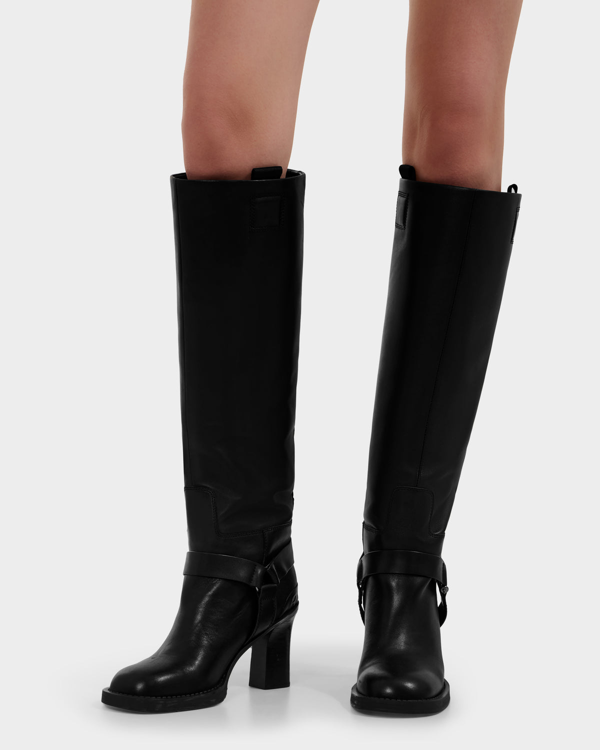 A person is wearing Rudsak ROSSEY - 8225574 Black knee-high leather boots with block heels and decorative straps, standing against a plain light background.