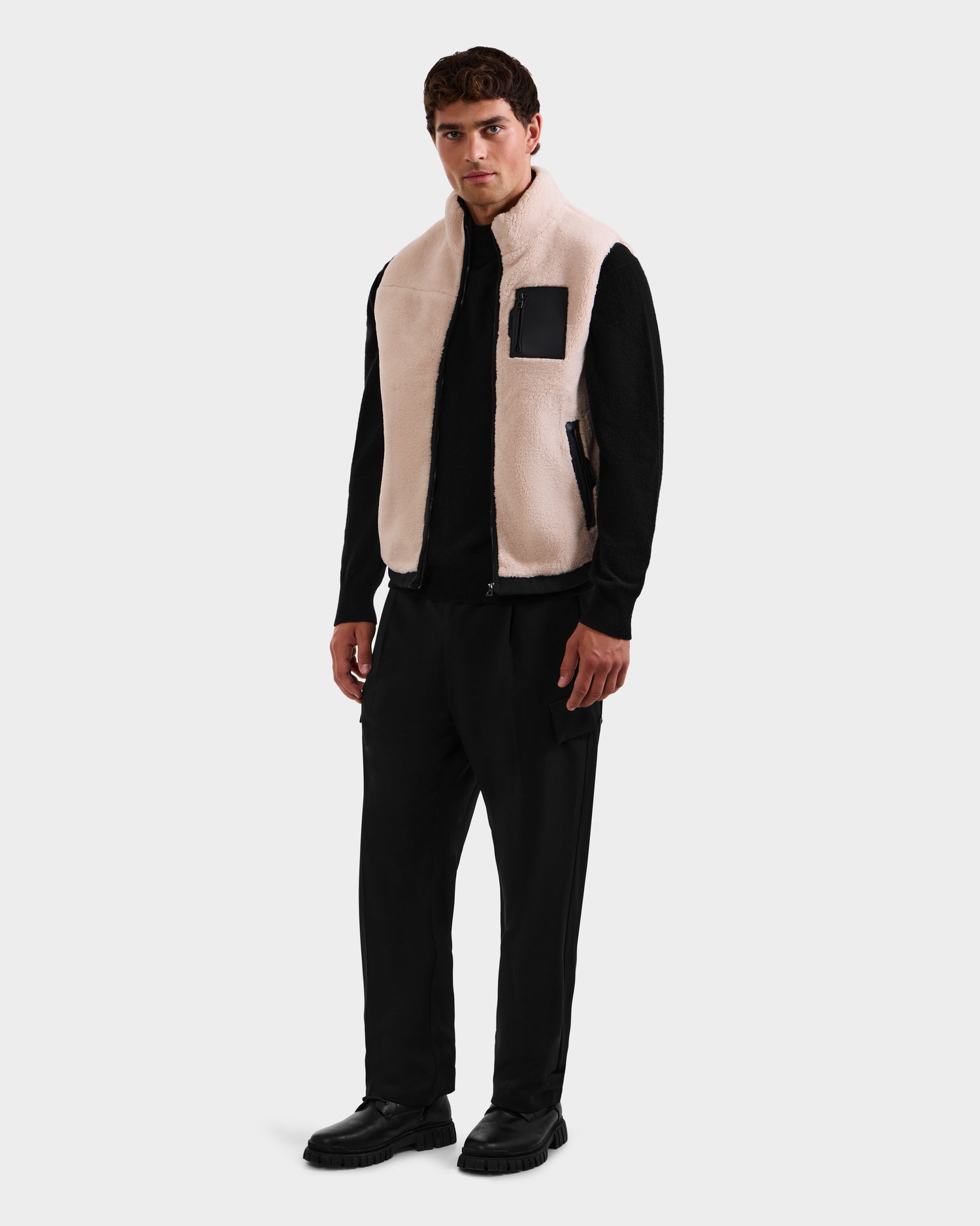 A man stands facing forward against a plain white background, wearing the Rudsak RUMMY - U524502 Vanilla M light beige polar fleece vest with a black chest pocket, over a black long-sleeve shirt, black pants, and black shoes.