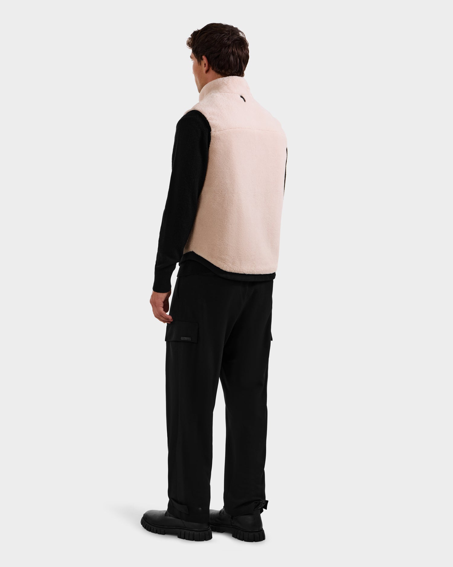 A man faces away, wearing the Rudsak RUMMY - U524502 Vanilla M polar fleece vest over a black long-sleeve shirt, black cargo pants, and black shoes, in front of a plain light background.