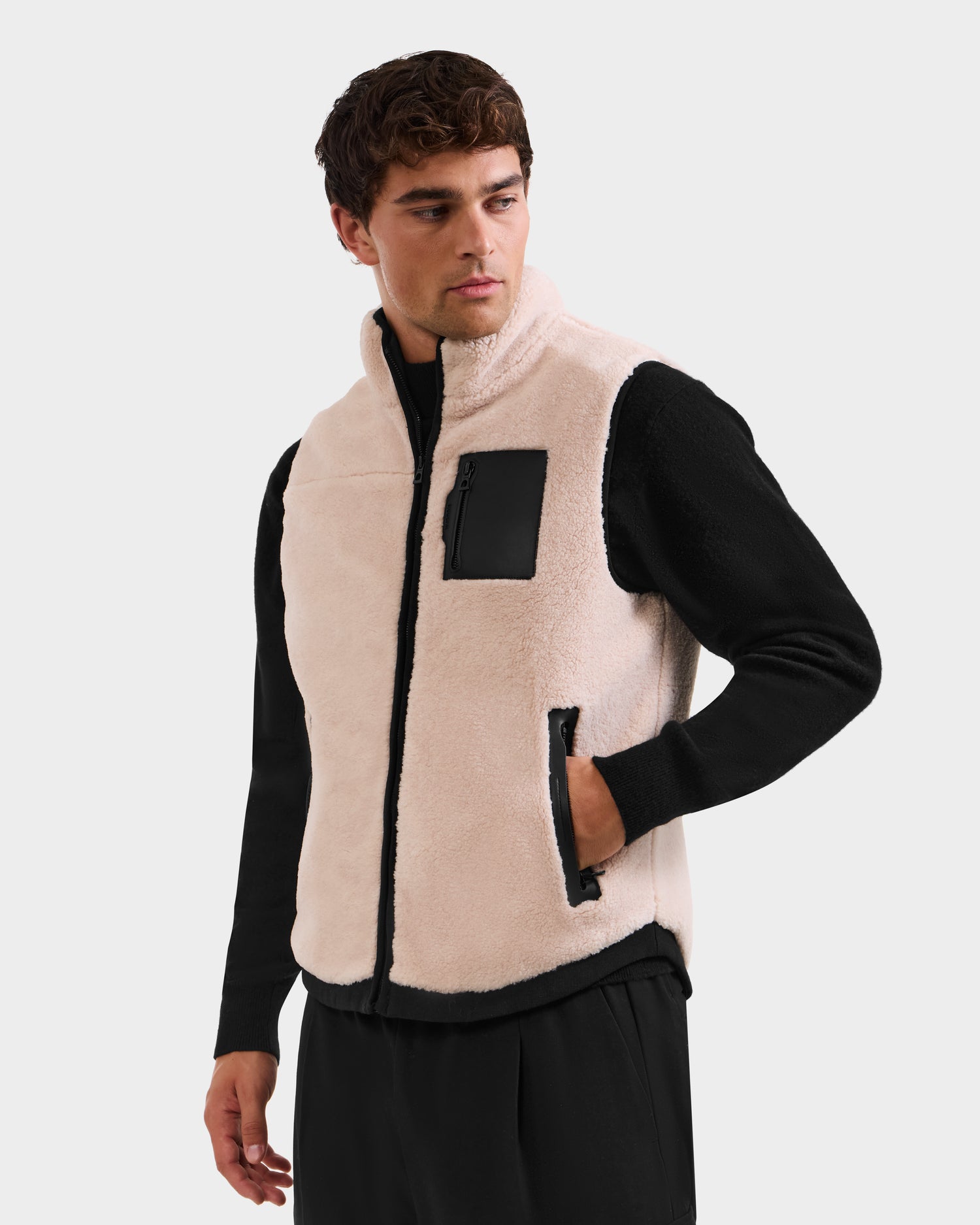 A man wears the Rudsak RUMMY - U524502 Vanilla M light beige polar fleece vest with black zipper pockets over a black long-sleeve shirt and black pants, standing against a plain light background.