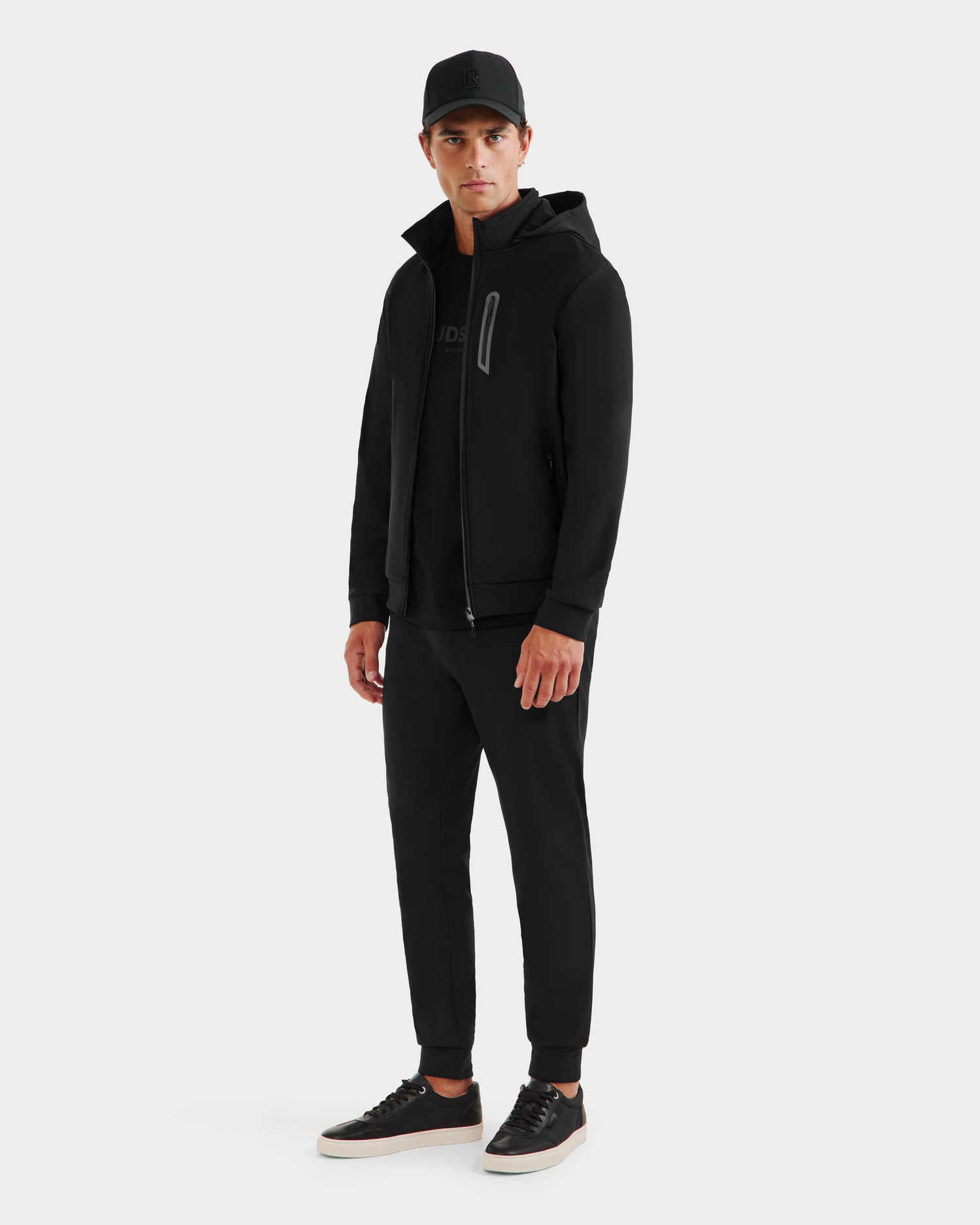 A man faces forward against a white background, wearing the Rudsak SAFRIN - 6525581 Black men’s sport jacket with a removable hood, paired with black jogger pants and black sneakers with white soles.