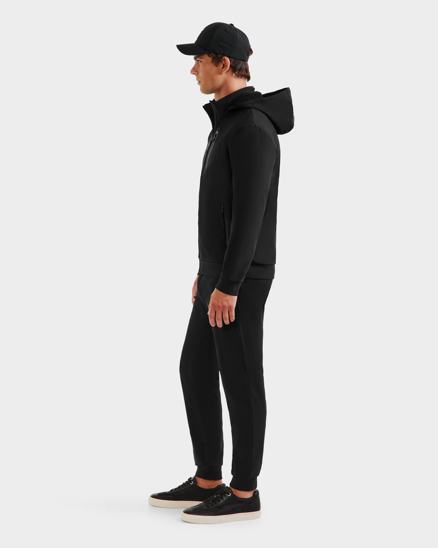 A man stands in profile against a white background, wearing the Rudsak SAFRIN - 6525581 Black men’s sport jacket with removable hood, paired with black pants and black sneakers with white soles.
