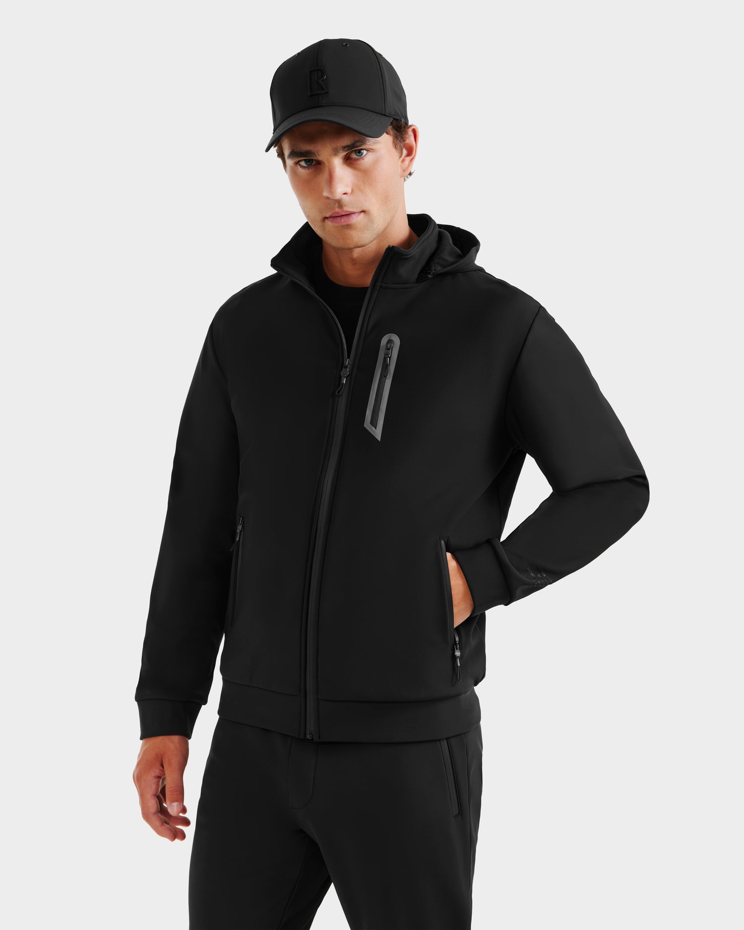 A man wearing the Rudsak SAFRIN - 6525581 Black men’s sport jacket, black pants, and a black baseball cap stands against a plain light gray background, looking at the camera with a neutral expression.