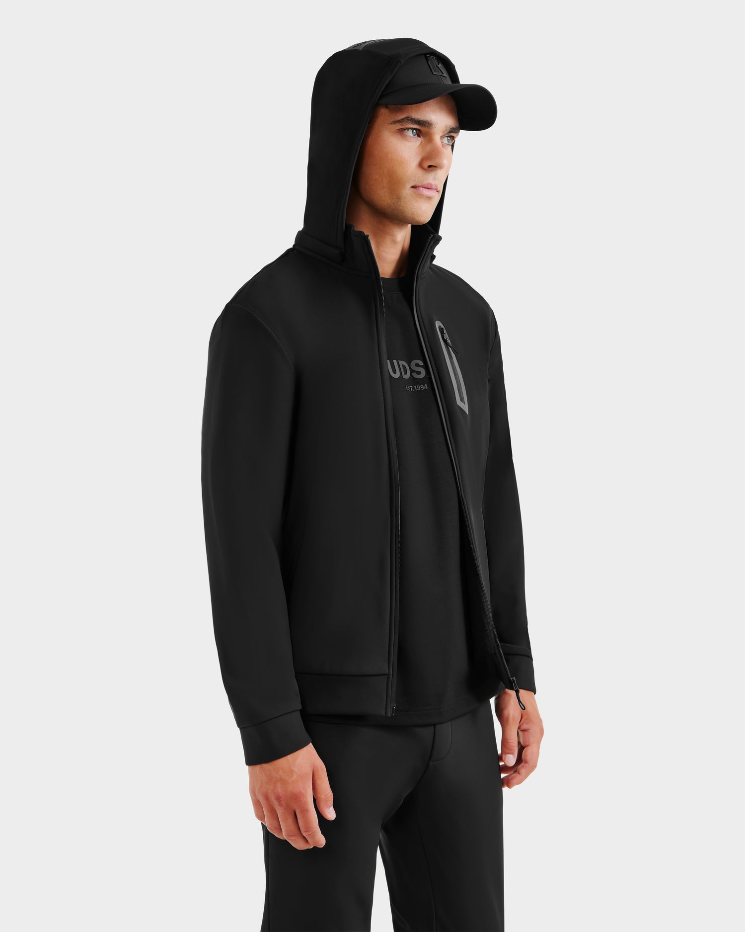 A man stands slightly turned against a plain light background, wearing the Rudsak SAFRIN - 6525581 Black men’s sport jacket with a removable hood, paired with a black cap, t-shirt, and pants.