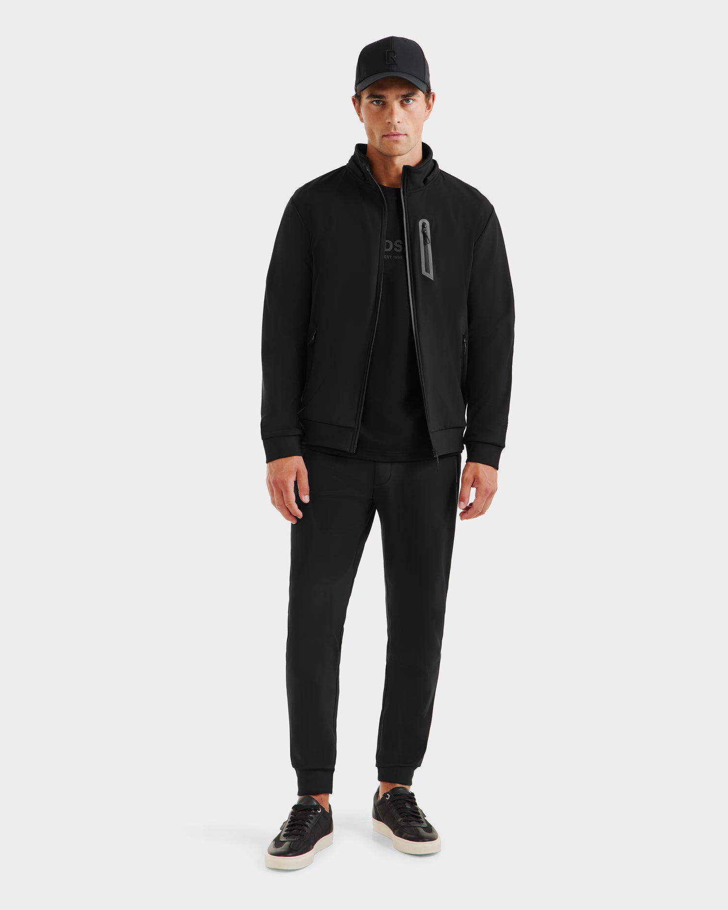 A man faces forward wearing the Rudsak SAFRIN - 6525581 Black mens sport jacket with a lightweight stretch shell and removable hood, paired with a black T-shirt, jogger pants, cap, and black sneakers with white soles against a light background.