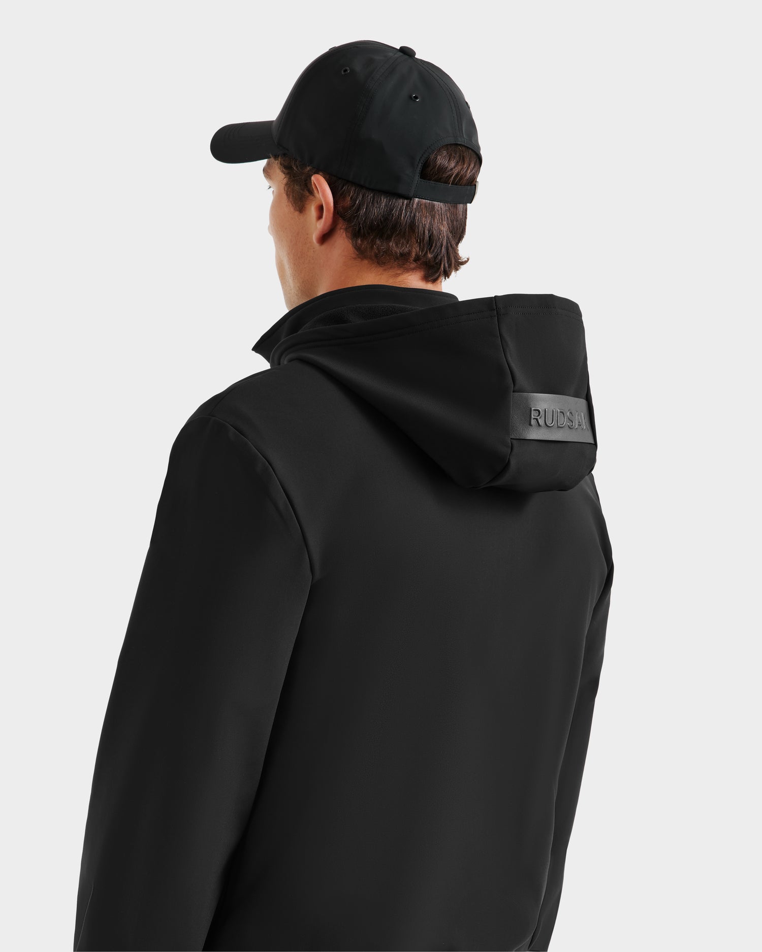 A person is seen from the back, wearing the Rudsak SAFRIN - 6525581 Black mens sport jacket with a removable hood, and a black baseball cap against a plain light background. The word RUDER appears on the hood.