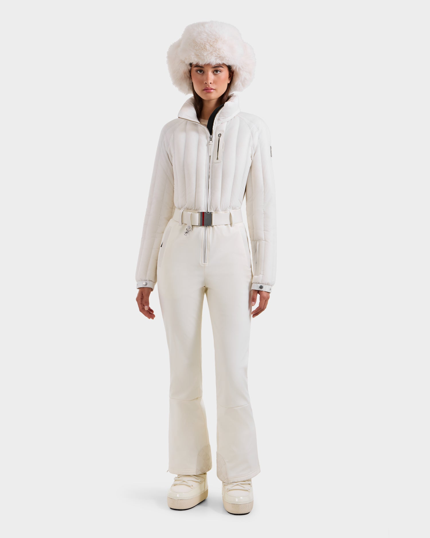 A person stands facing forward in the Rudsak SAGE - 8124782 Iceland all-white winter outfit, featuring a puffy jacket, flared pants, platform boots, a belt, and a large fluffy hat, against a plain white background.