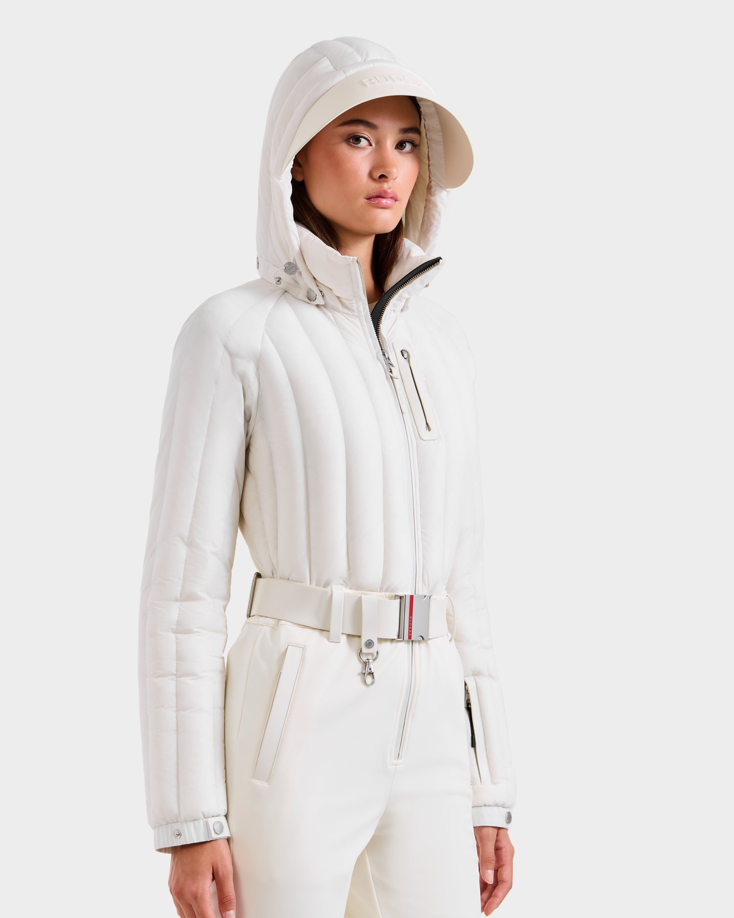 A woman wears the Rudsak SAGE - 8124782 Iceland women’s ski suit, featuring a stylish fitted white design with hood, high-waisted pants, and wide belt—blending fashion and technical performance against a white background.