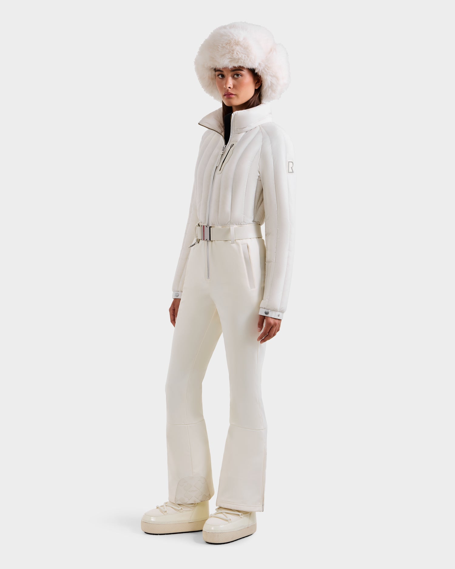 A woman models the Rudsak SAGE - 8124782 Iceland all-white ski suit with technical features and a belted design, paired with a fluffy white hat and platform boots, set against a plain white background.
