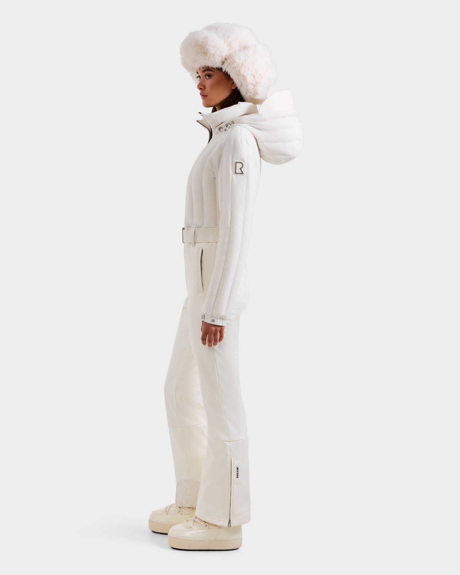 A woman in profile wears the Rudsak SAGE - 8124782 Iceland ski suit: an all-white puffy jacket with hood, belted pants, chunky boots, and a large fur hat—an ensemble designed for technical performance against a plain background.