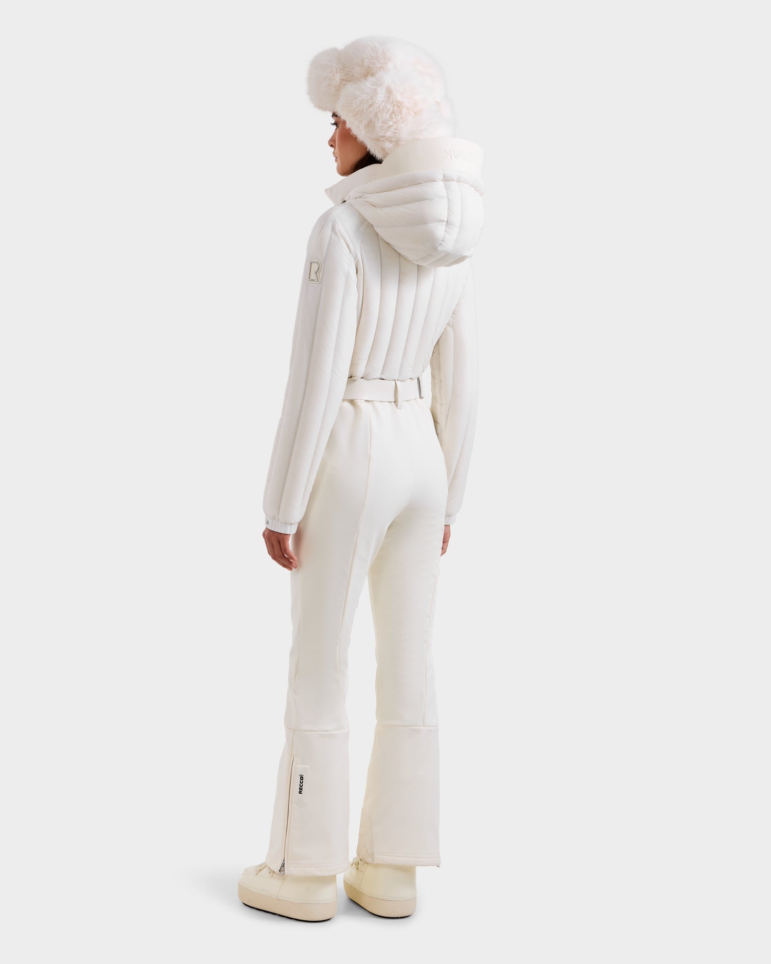 A person stands facing away, wearing the SAGE - 8124782 Iceland all-white women’s ski suit by Rudsak, featuring a hooded padded jacket with RECCO rescue tech, fitted pants, boots, and a fluffy hat against a plain light background.