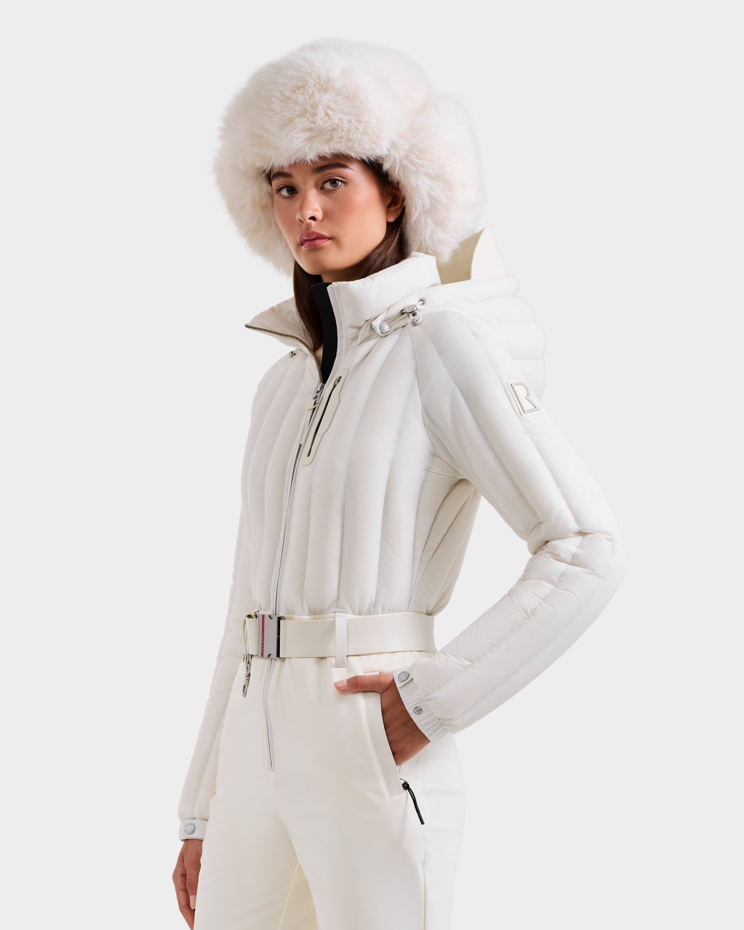 A woman models the Rudsak SAGE - 8124782 Iceland white winter jacket with belt, matching pants, and a fluffy fur hat, confidently highlighting its high-end ski suit-inspired technical performance.